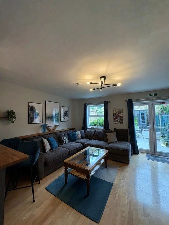 Modern living room with dark sectional sofa, glass coffee table, blue rug, framed wall art, and glass patio doors