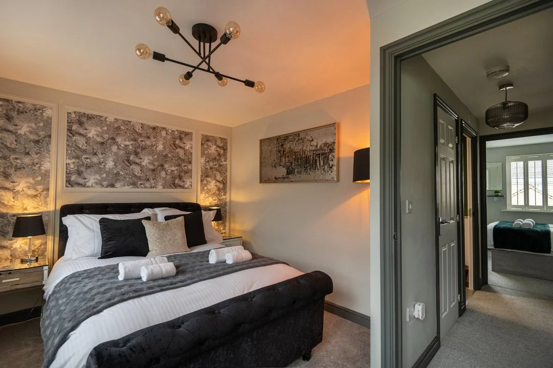 Elegant modern bedroom with a black upholstered bed, white linens, abstract wall art, and a contemporary chandelier, overlooking a hallway and adjoining bedroom.