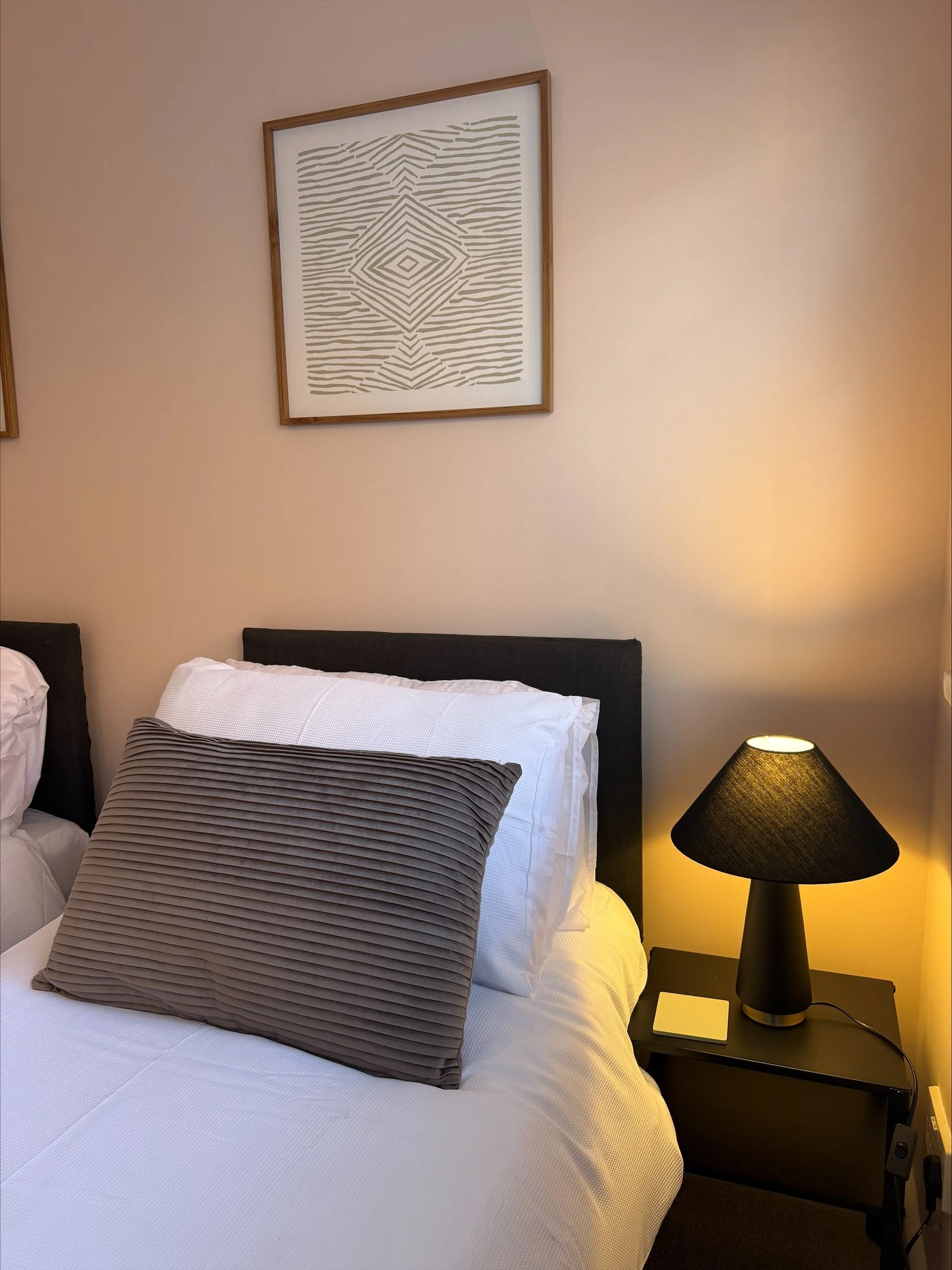 Cozy bedroom with a neatly made white bed, gray ribbed accent pillow, black nightstand and lamp, and geometric framed wall art.