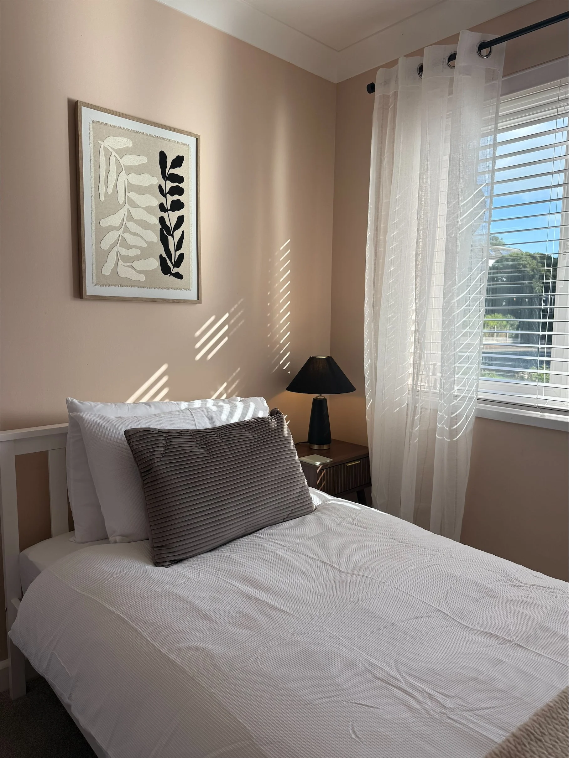 Sunlit bedroom with a neatly made white bed, gray accent pillow, black bedside lamp, botanical wall art, and sheer curtains.