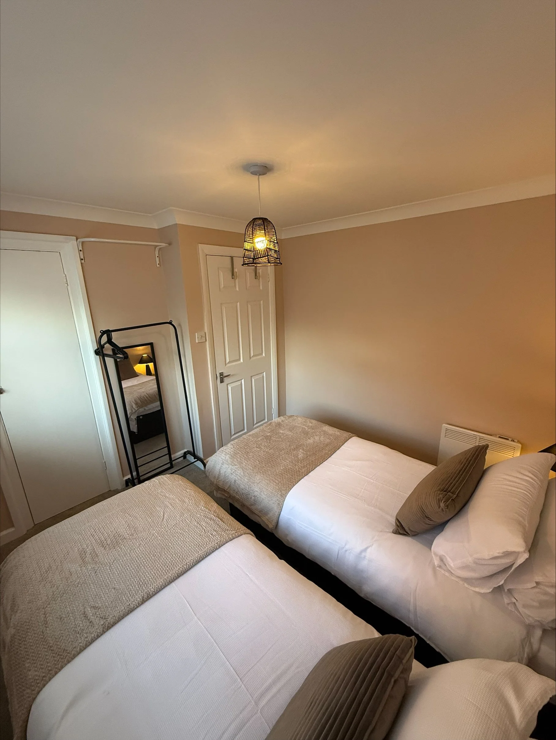 Cozy bedroom with two twin beds, white linens, beige throws, a woven pendant light, and a full-length mirror