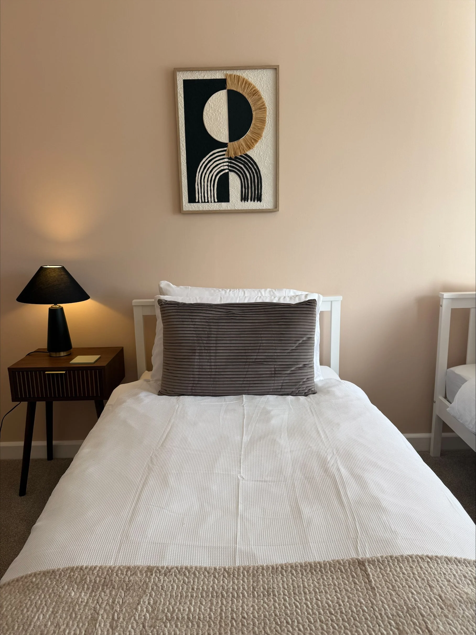 Cozy bedroom with a neatly made white bed, gray accent pillow, black bedside lamp, and framed geometric wall art.