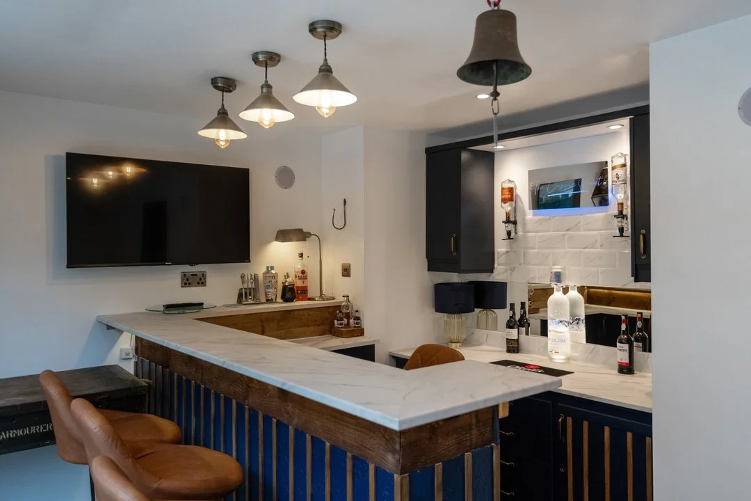 Modern home bar with marble countertop, blue cabinets, leather stools, wall-mounted TV, pendant lights, and liquor bottles