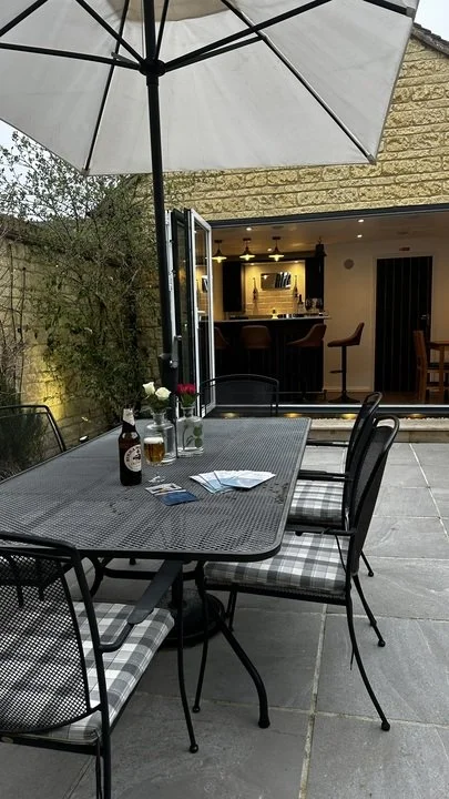 Outdoor patio dining table with umbrella, chairs, drinks, flowers, and an open doorway to a warmly lit bar.