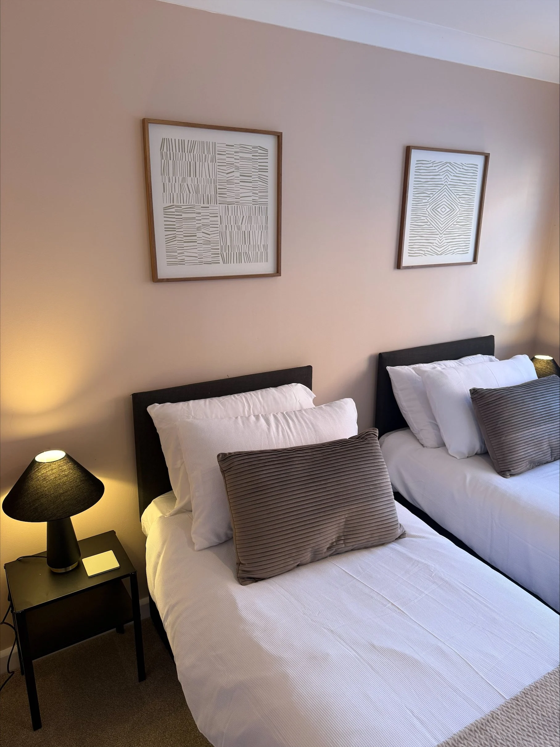 Cozy bedroom with two neatly made beds, gray accent pillows, black bedside table lamps, and framed abstract wall art