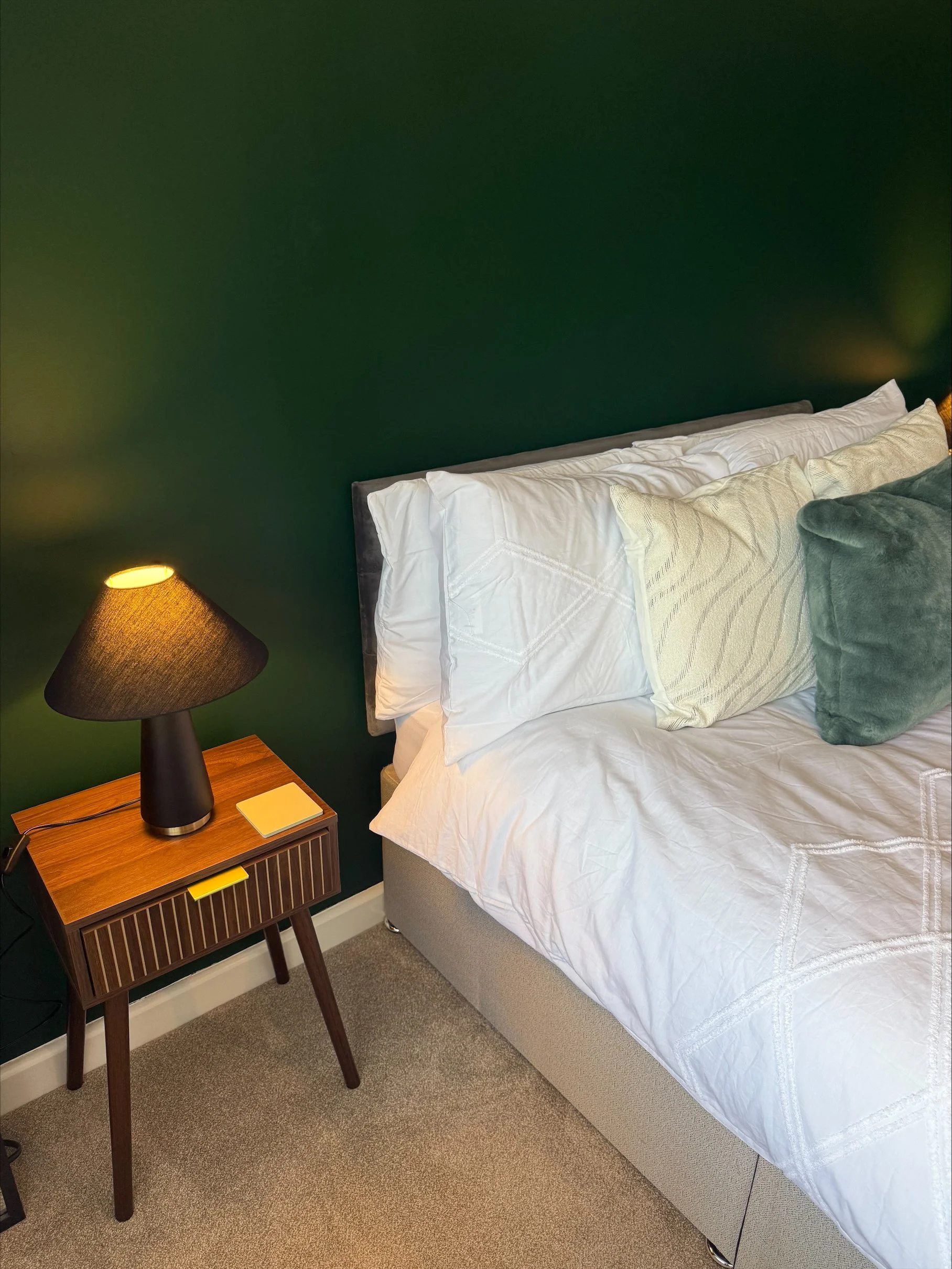 Cozy bedroom with a white bed, textured pillows, dark green wall, and midcentury wooden nightstand with a lit table lamp
