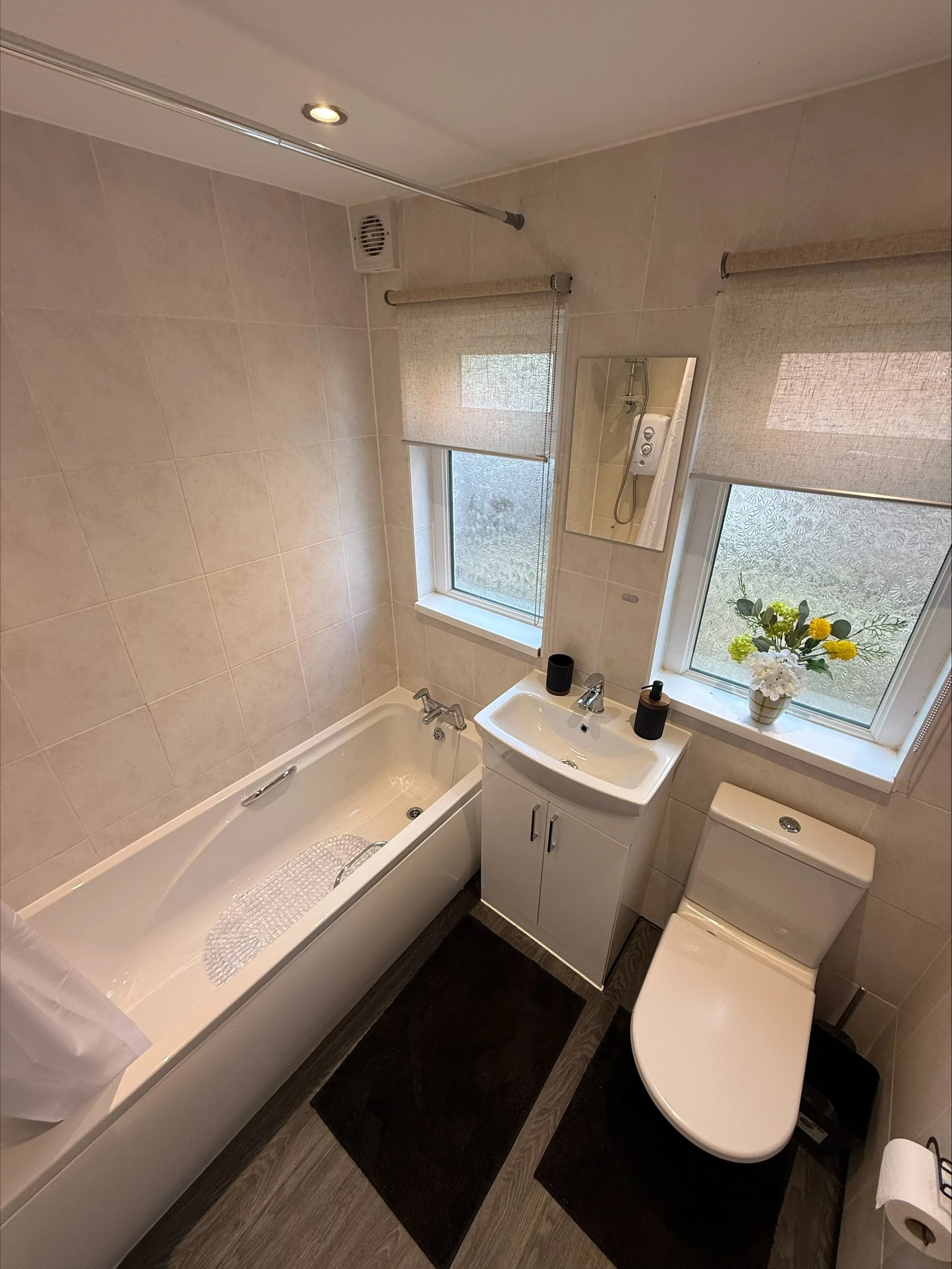 Bright tiled bathroom with bathtub, sink vanity, toilet, frosted windows, roller blinds, and floral decor