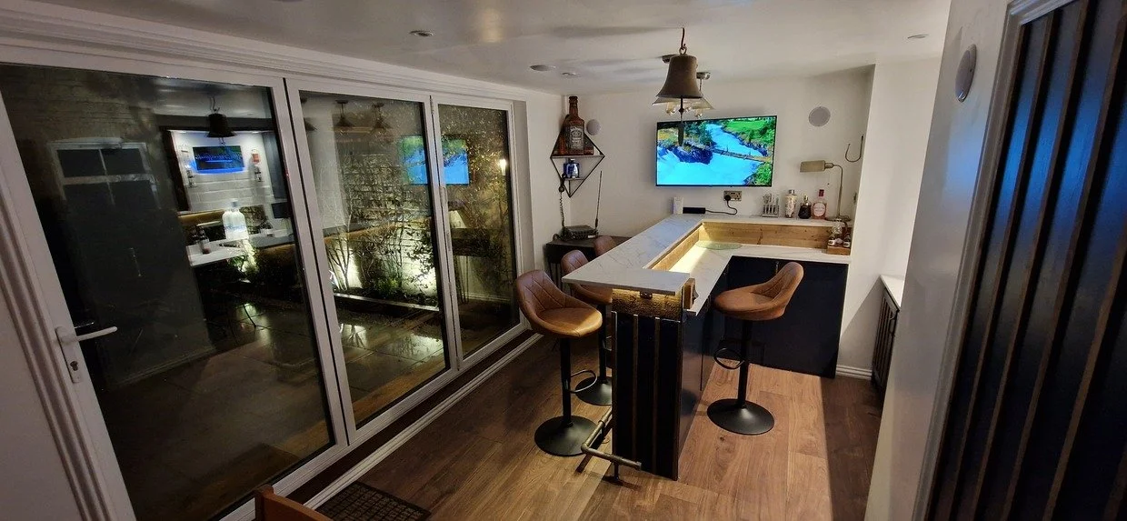 Modern home bar with marble countertop, leather stools, wall-mounted TV, glass doors, and indoor garden view