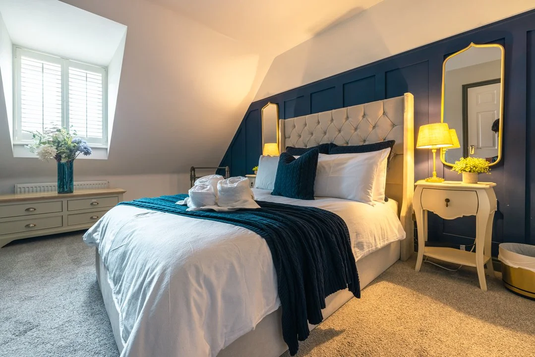 Elegant bedroom with a tufted bed, navy accent wall, gold-trimmed mirrors, bedside lamp, and floral vase
