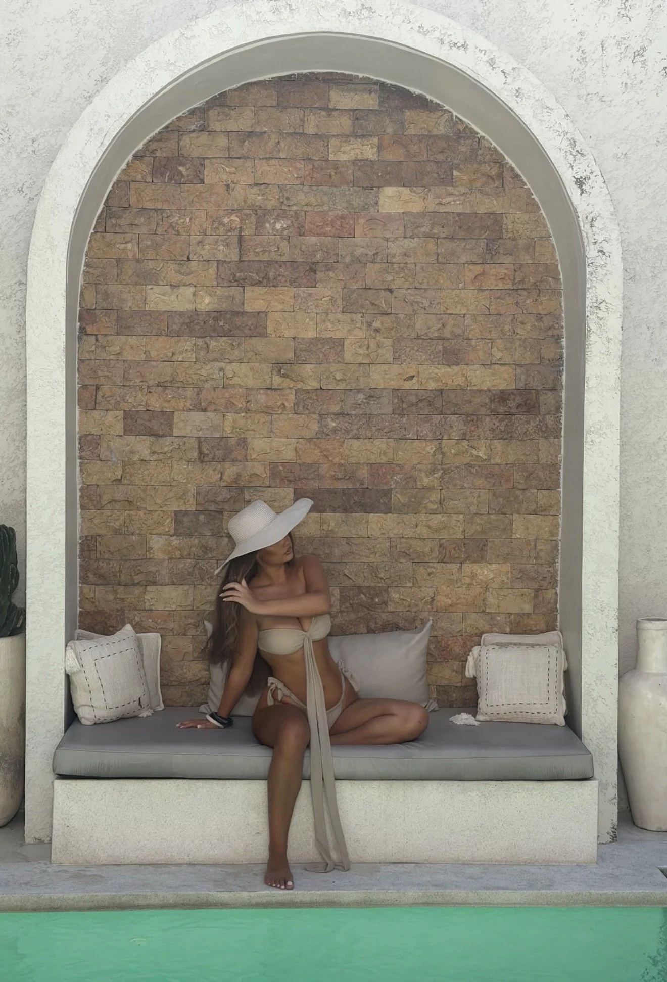 A woman in a beige bikini and large sun hat sitting in a built-in outdoor seating area with beige pillows, a brick wall backdrop, and square white decorative objects beside her.