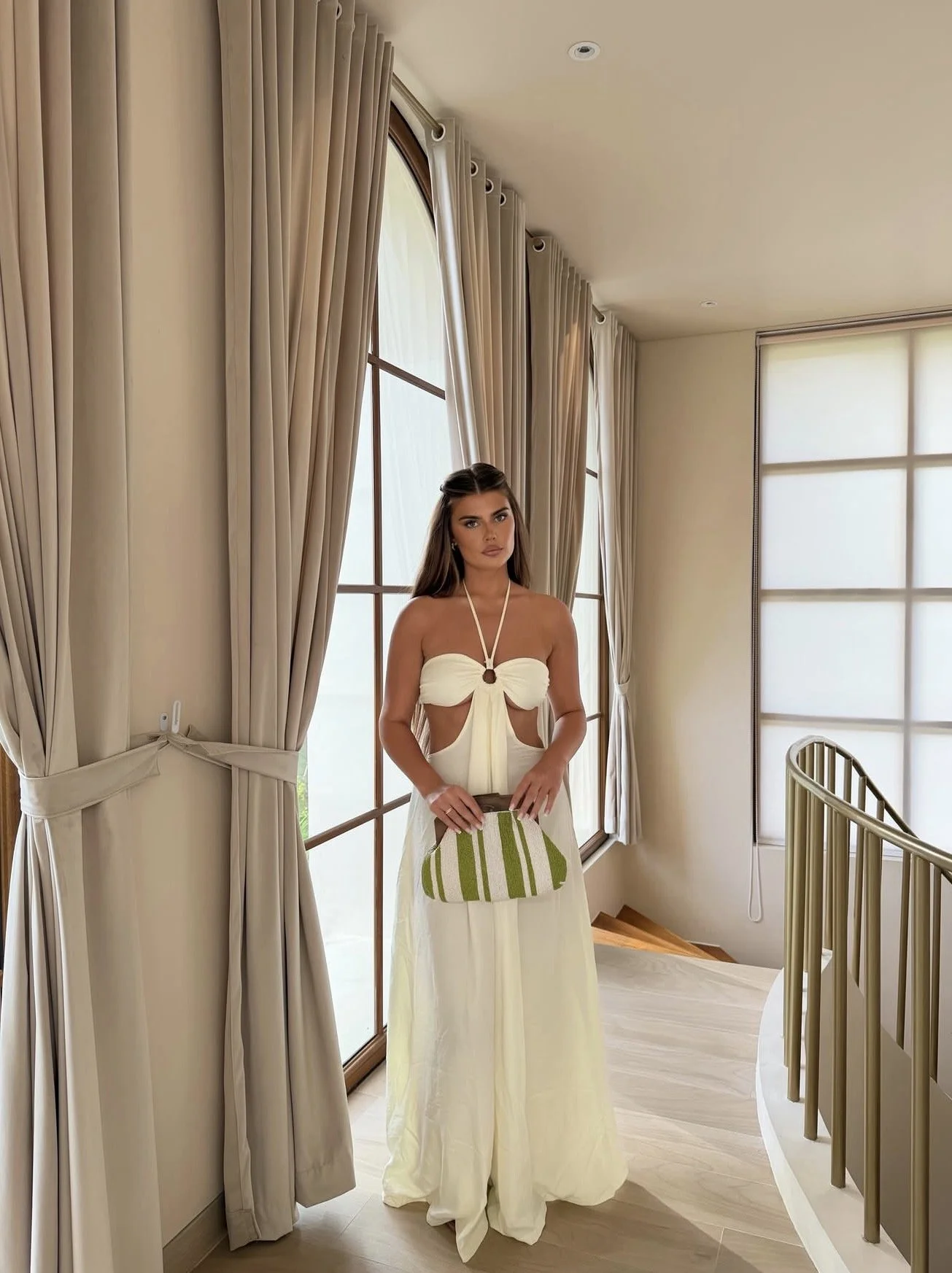 A woman in a white dress standing in a room with large window curtains, holding a striped green and white purse, near a staircase railing.