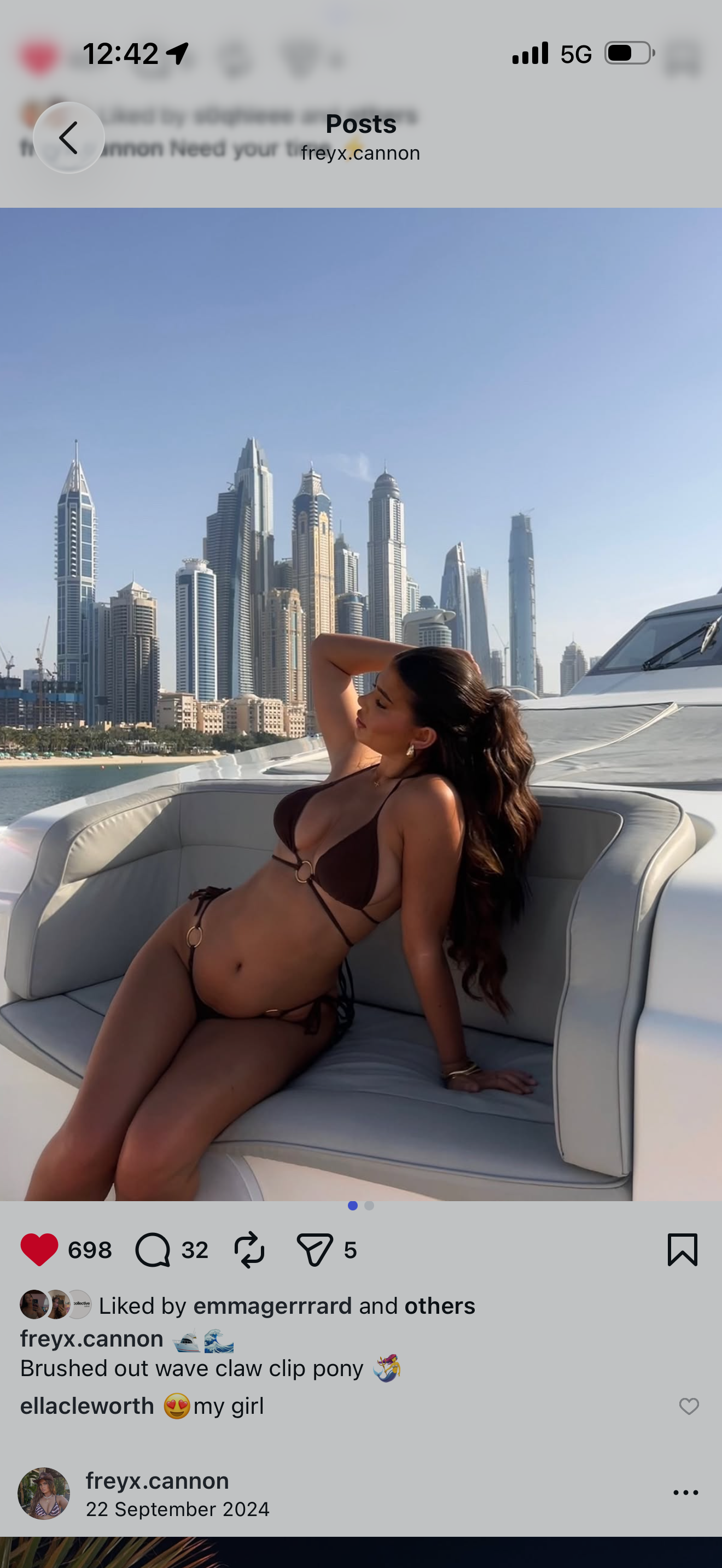 A woman in a brown bikini is sitting on a white yacht with a city skyline of tall buildings in the background, under a clear blue sky.