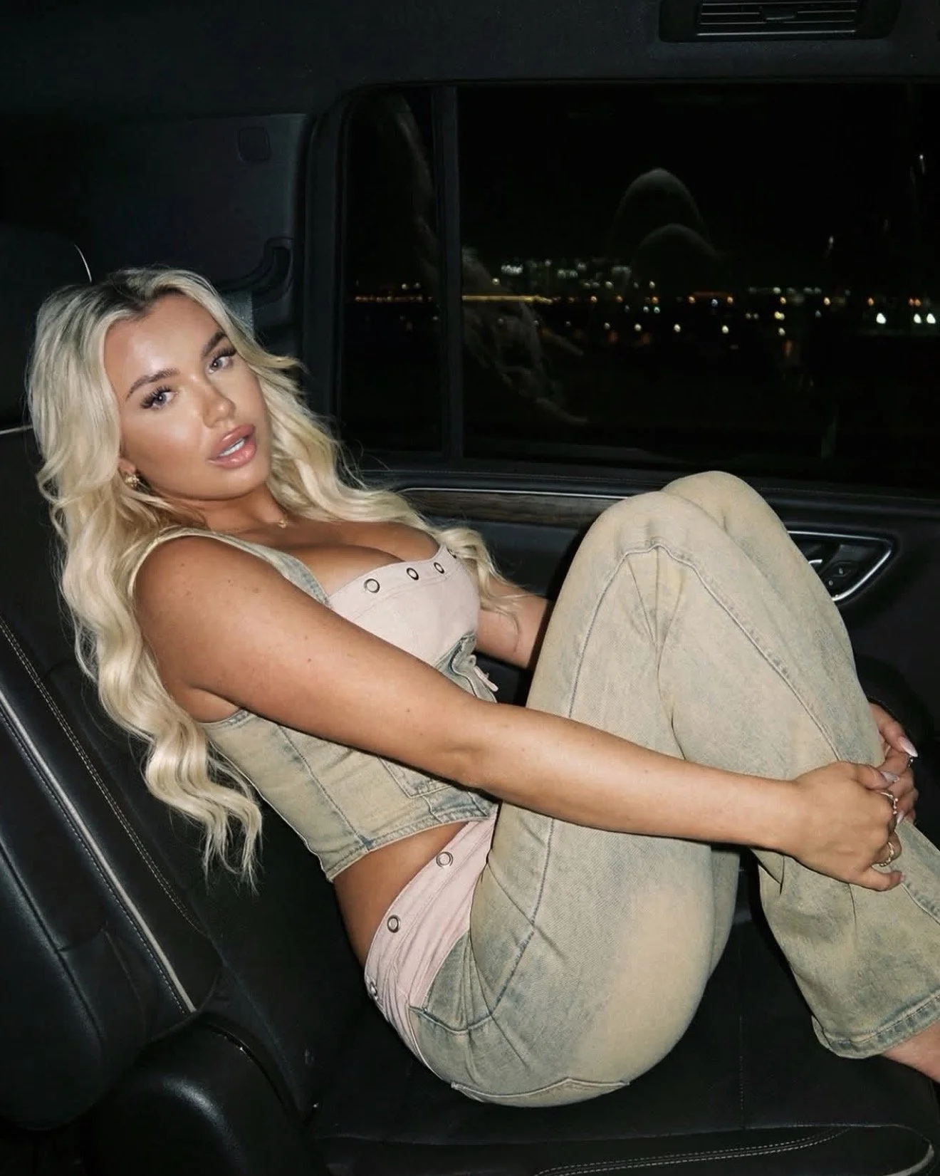 A woman with long blonde hair reclining inside a car at night, looking at the camera, with city lights visible through the window behind her.