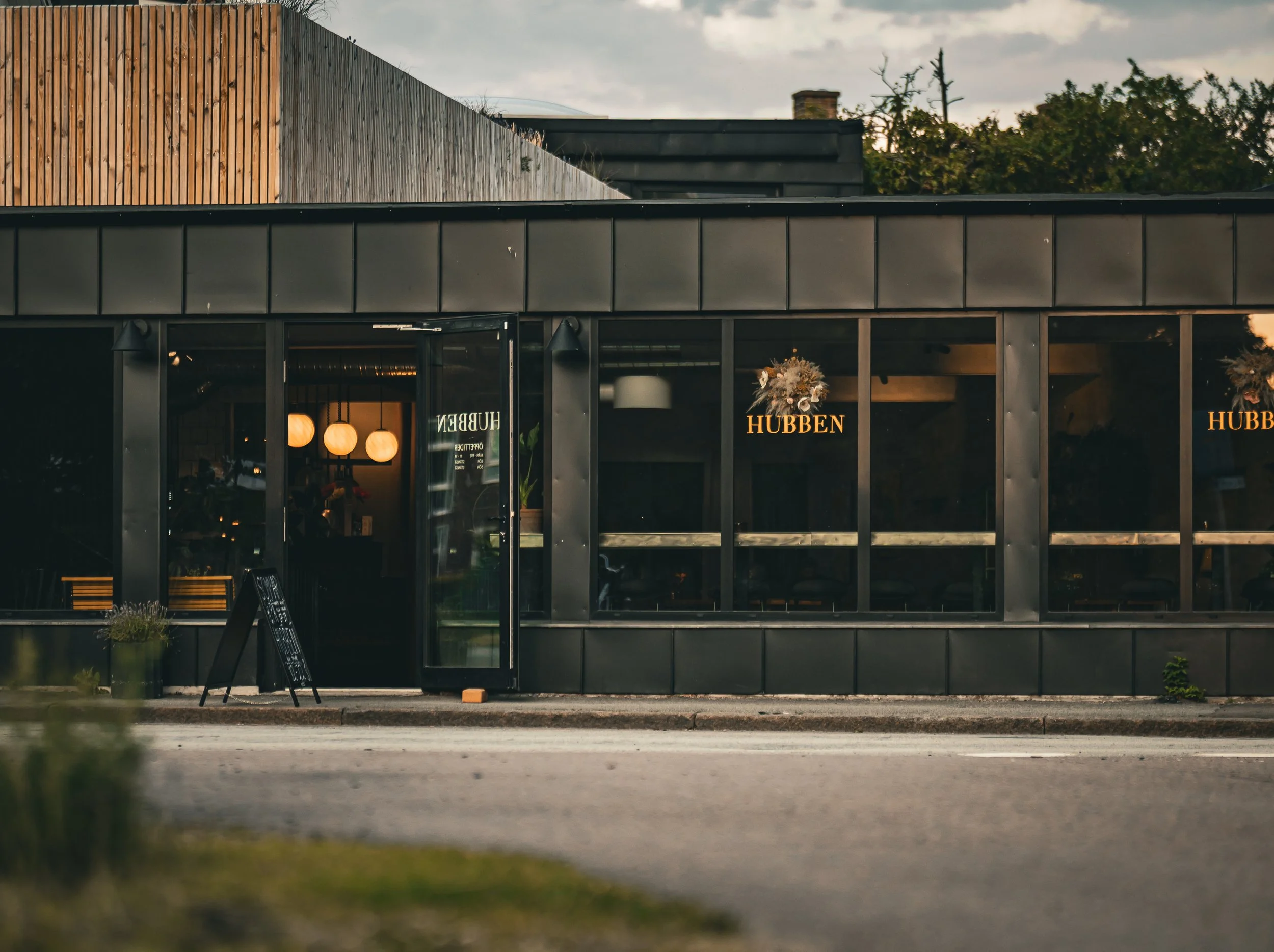 En modern byggnad med stora glasfönster och svarta väggar, inredning som syns genom fönstren inkluderar ljuslampor och blommor, utomhusmiljö med en gata framför och några träd i bakgrunden.
