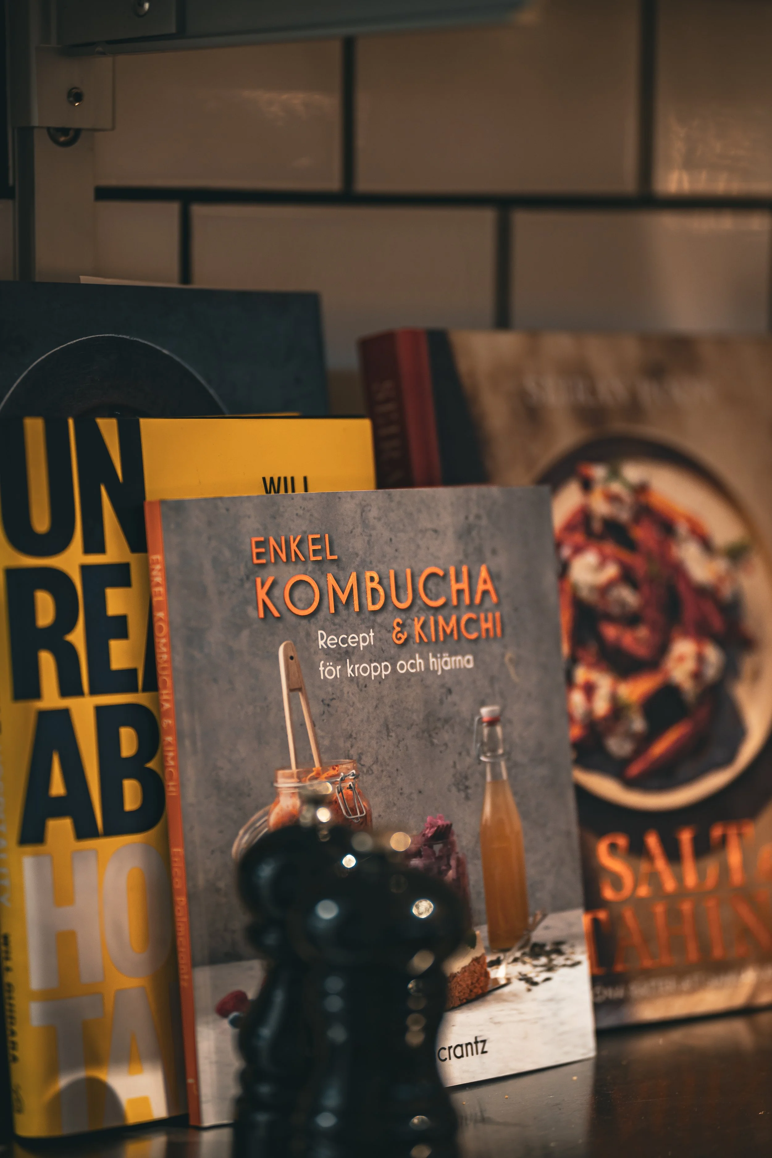 Böcker om hälsa och mat, bland annat om kombucha och kimchi, visas på ett hyllplan, med en pizza i bakgrunden.