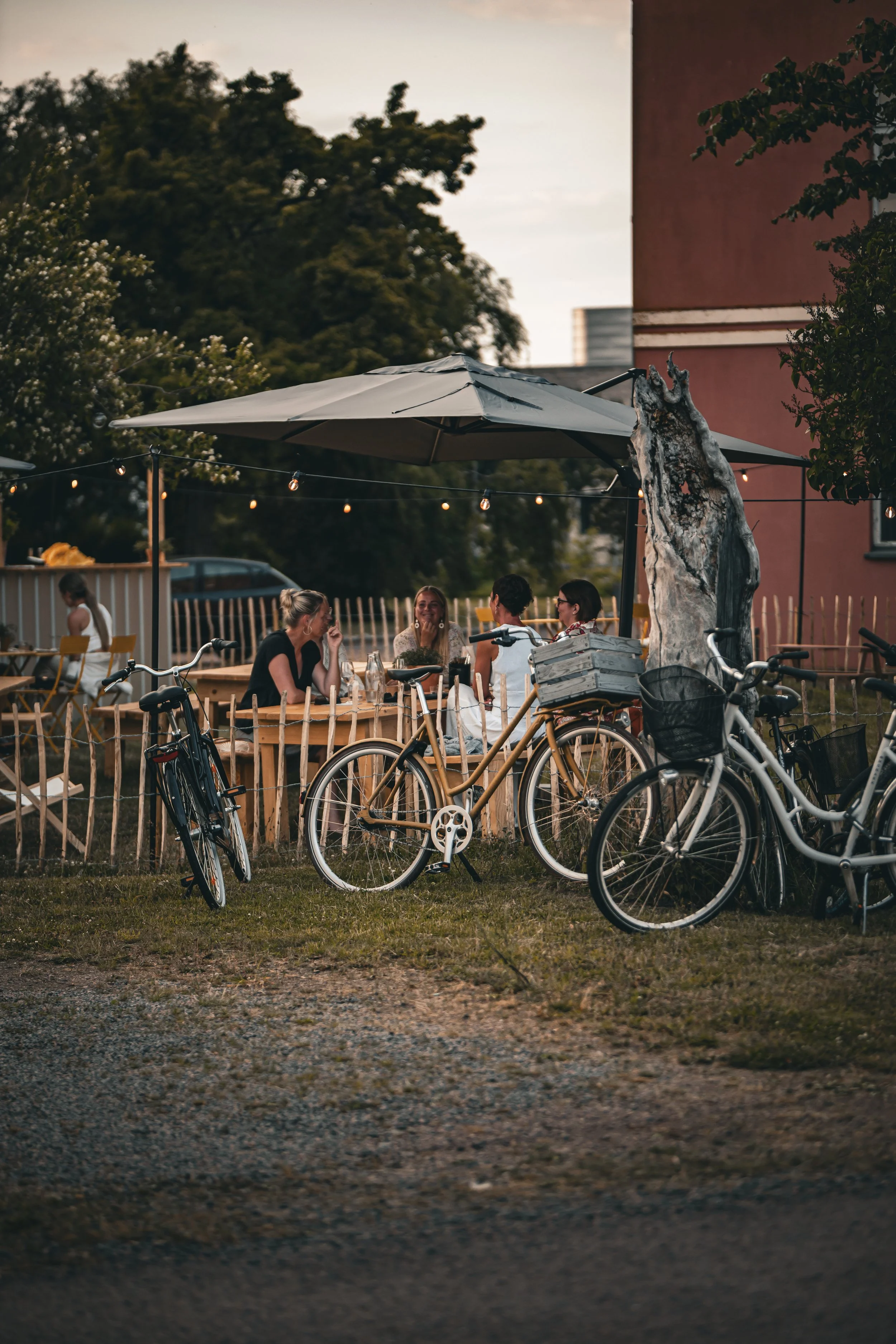 Grupper av människor som äter och pratar vid ett bord utomhus på en uteservering under kvällen, med cyklar parkerade framför.