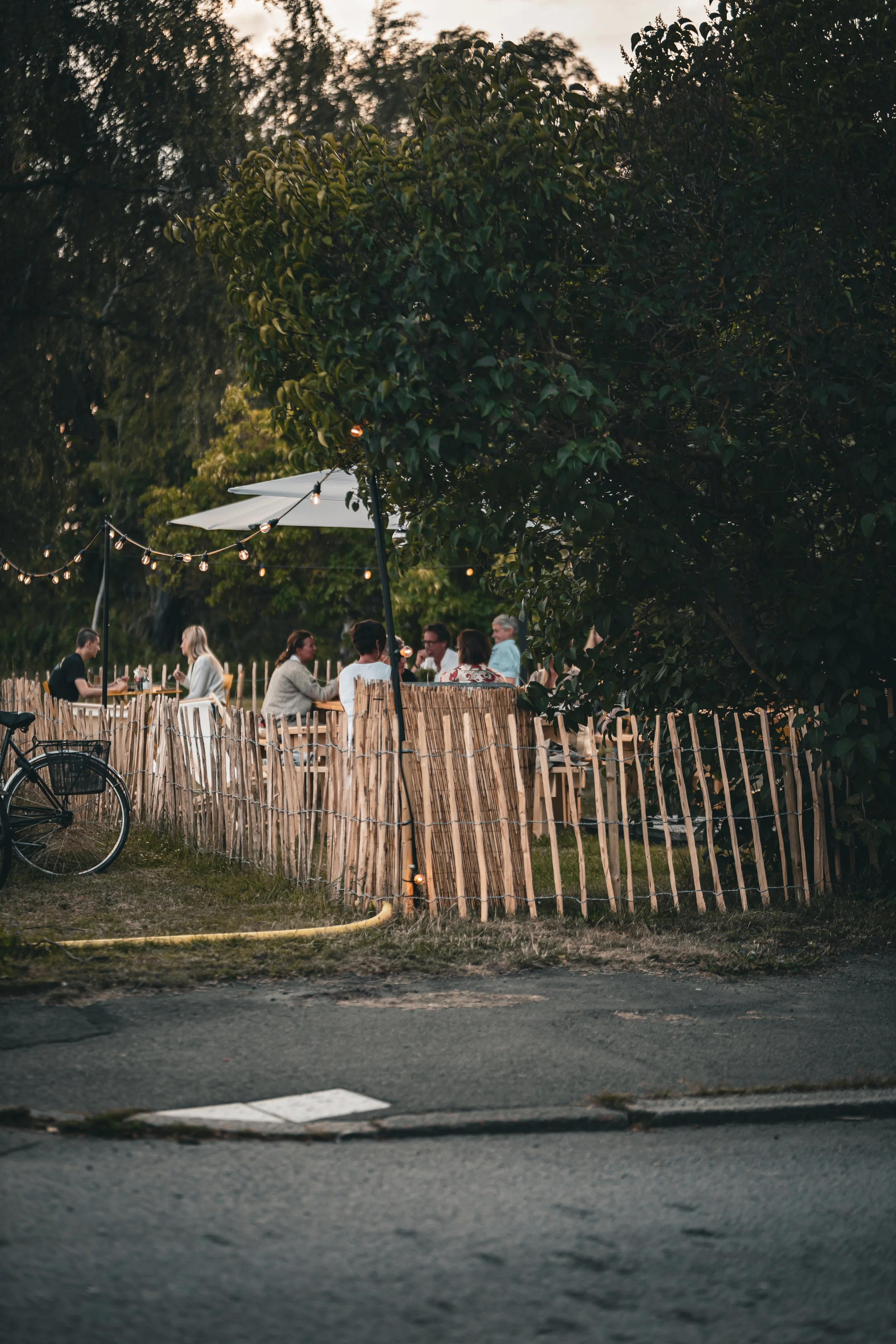 Folk som äter och socialiserar i en utomhusmiljö under svagt ljus, delvis skymningsljus, med träd och ett stängsel av trä runt området.