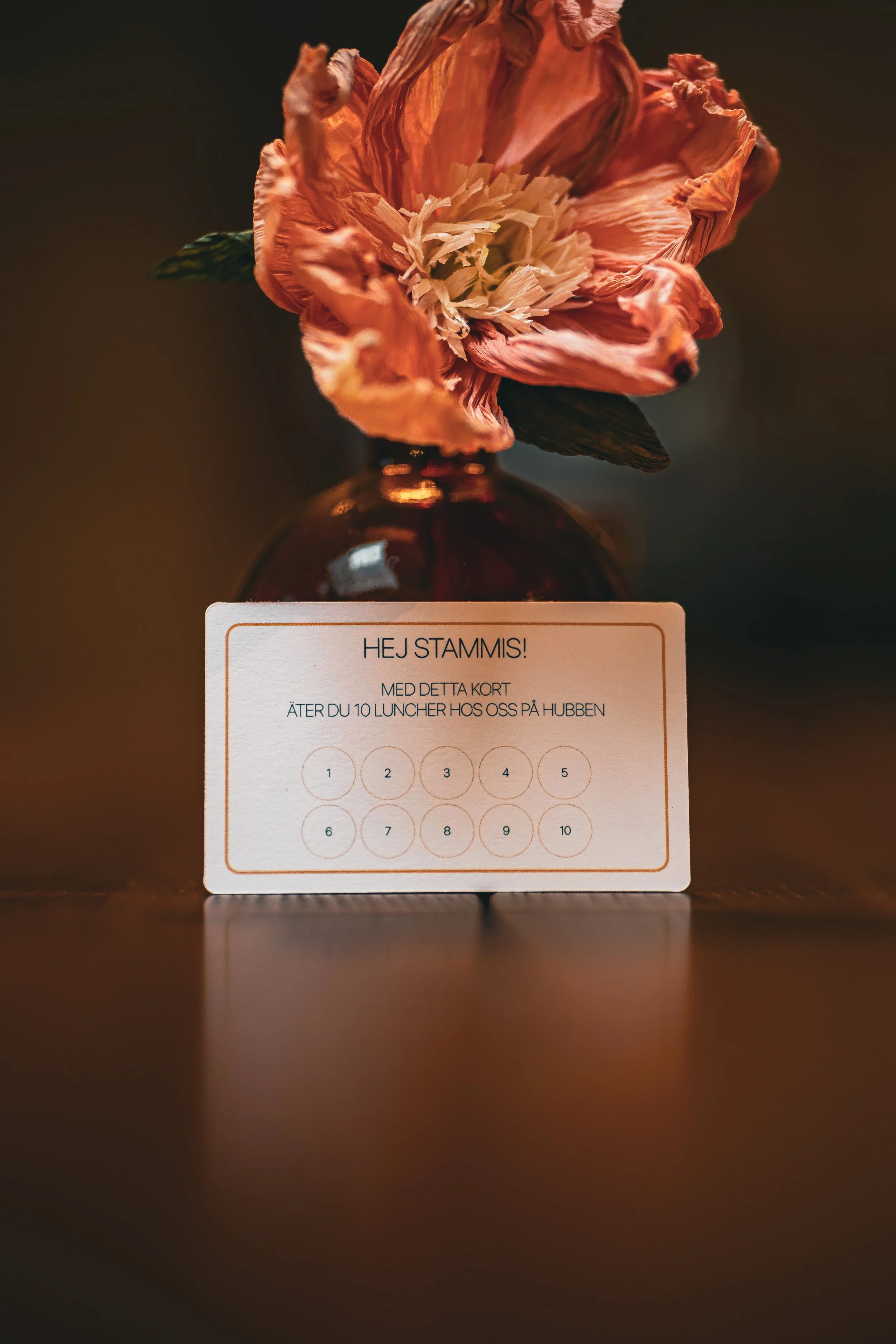 Ett blomarrangemang i en liten glassflaska med ett kort som säger ’HEJ STAMMIS!’ och en lapp med text om lunch på ett bord.