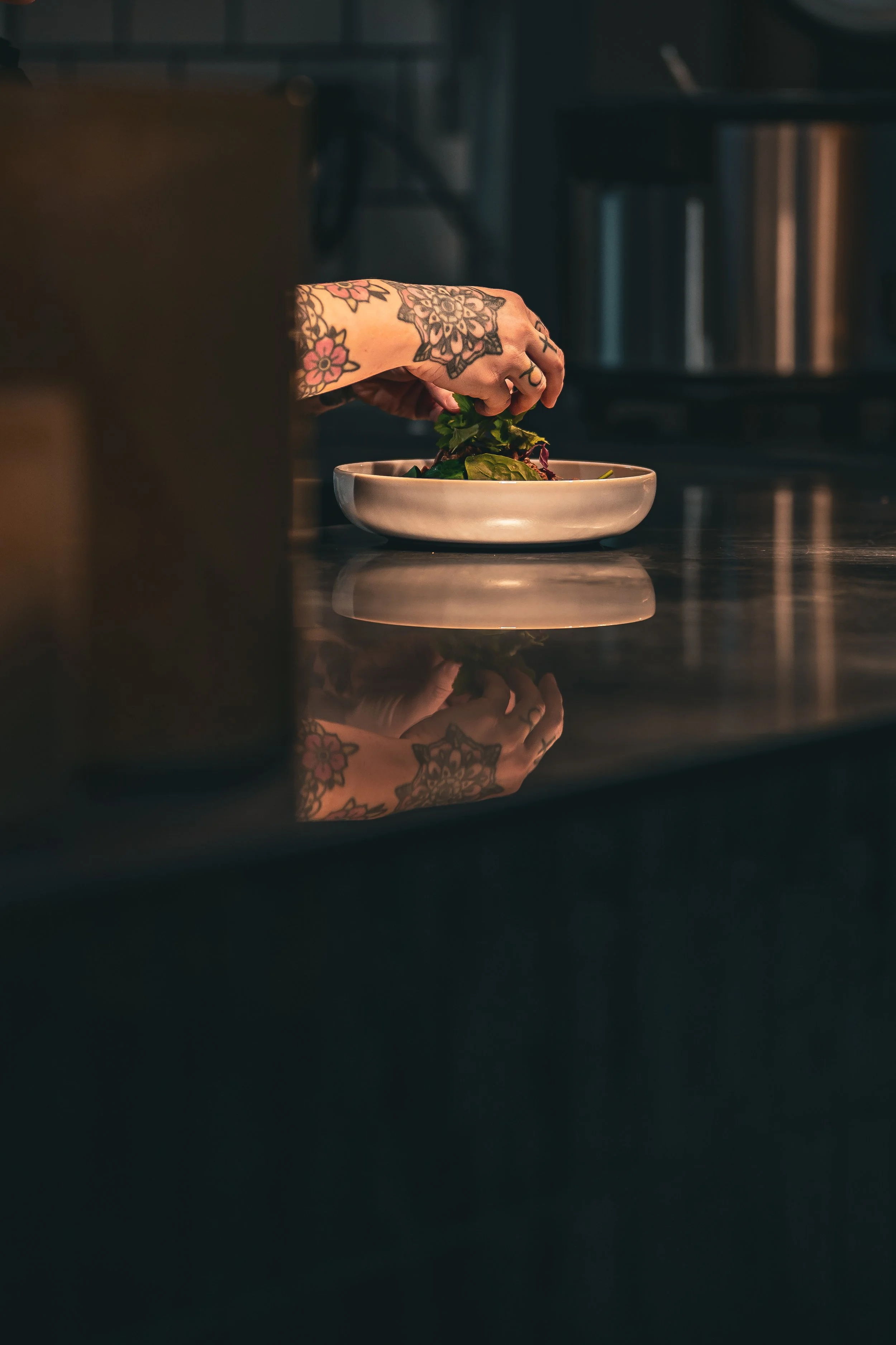 En tatueringad arm som placerar en hand i en skål med sallad på ett mörkt köksbänk.