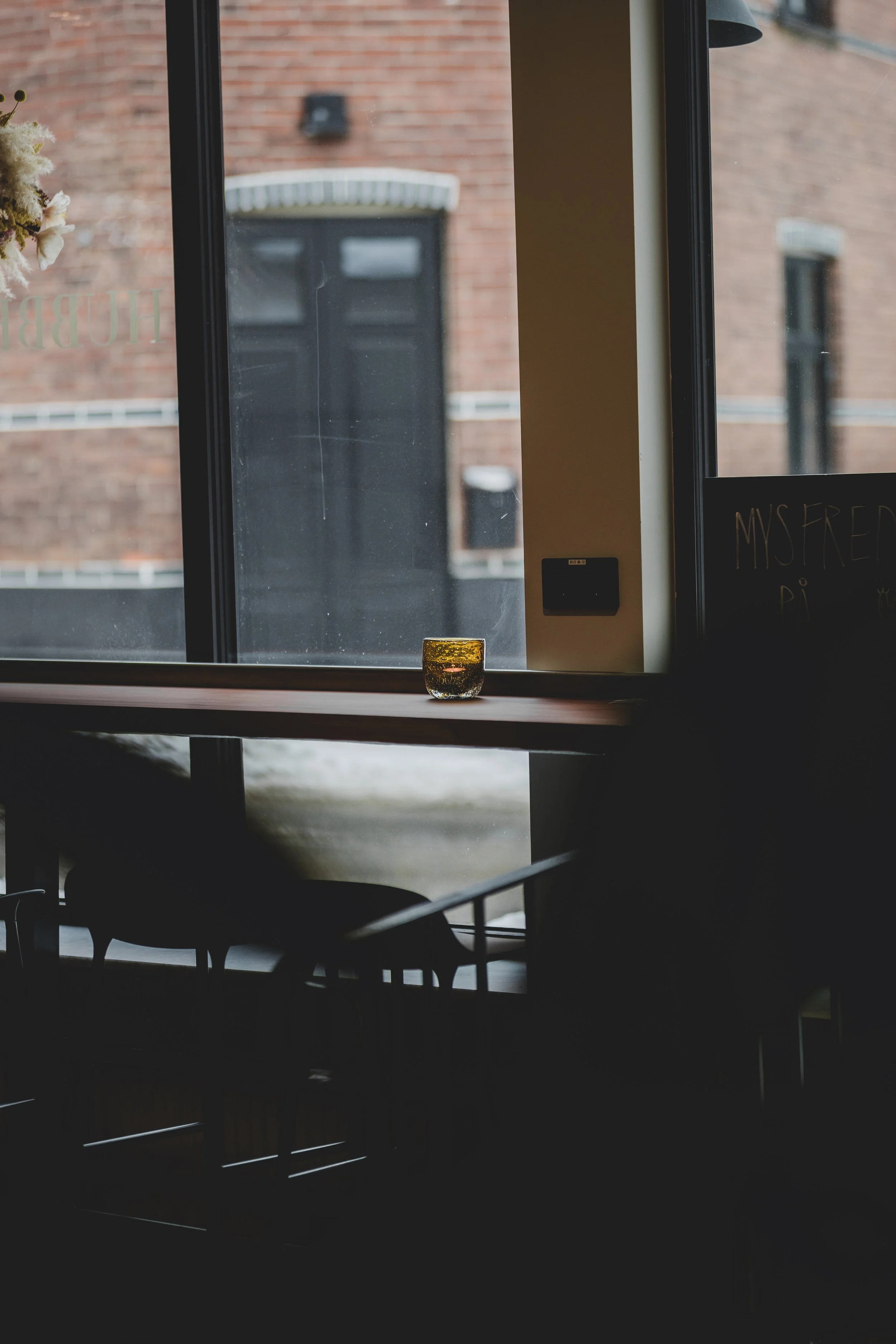 Ett fönster mot en fasadvägg. En liten doftljus eller glas är placerad på fönsterbrädan. Utsidan visar en röd tegelvägg och en fönster med vit taklist.
