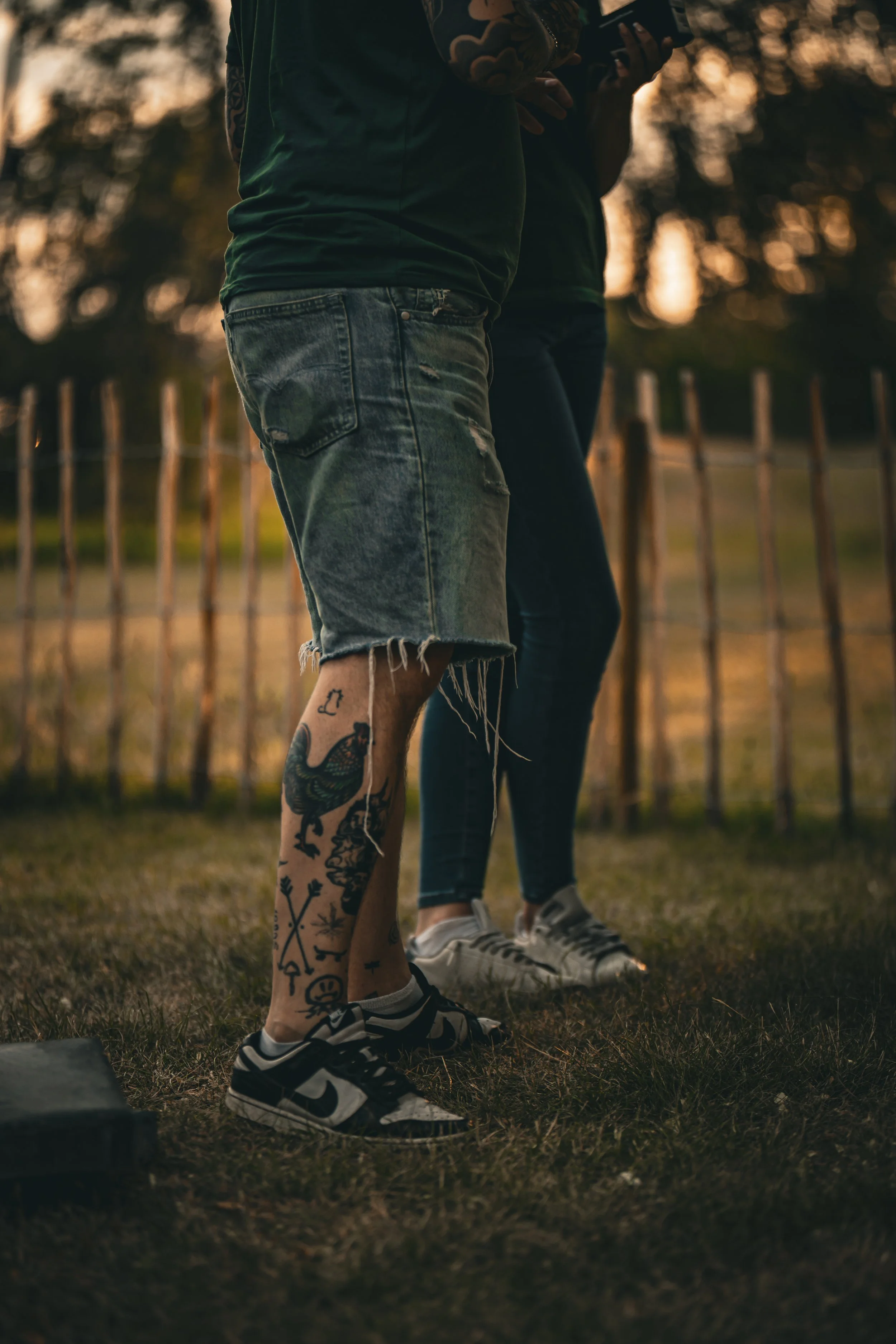 Två personer står utomhus i skymningen, den ena med tatueringar på vaden och den andra med jeans och vita sneakers, omgivna av ett trästaket och träd i bakgrunden.