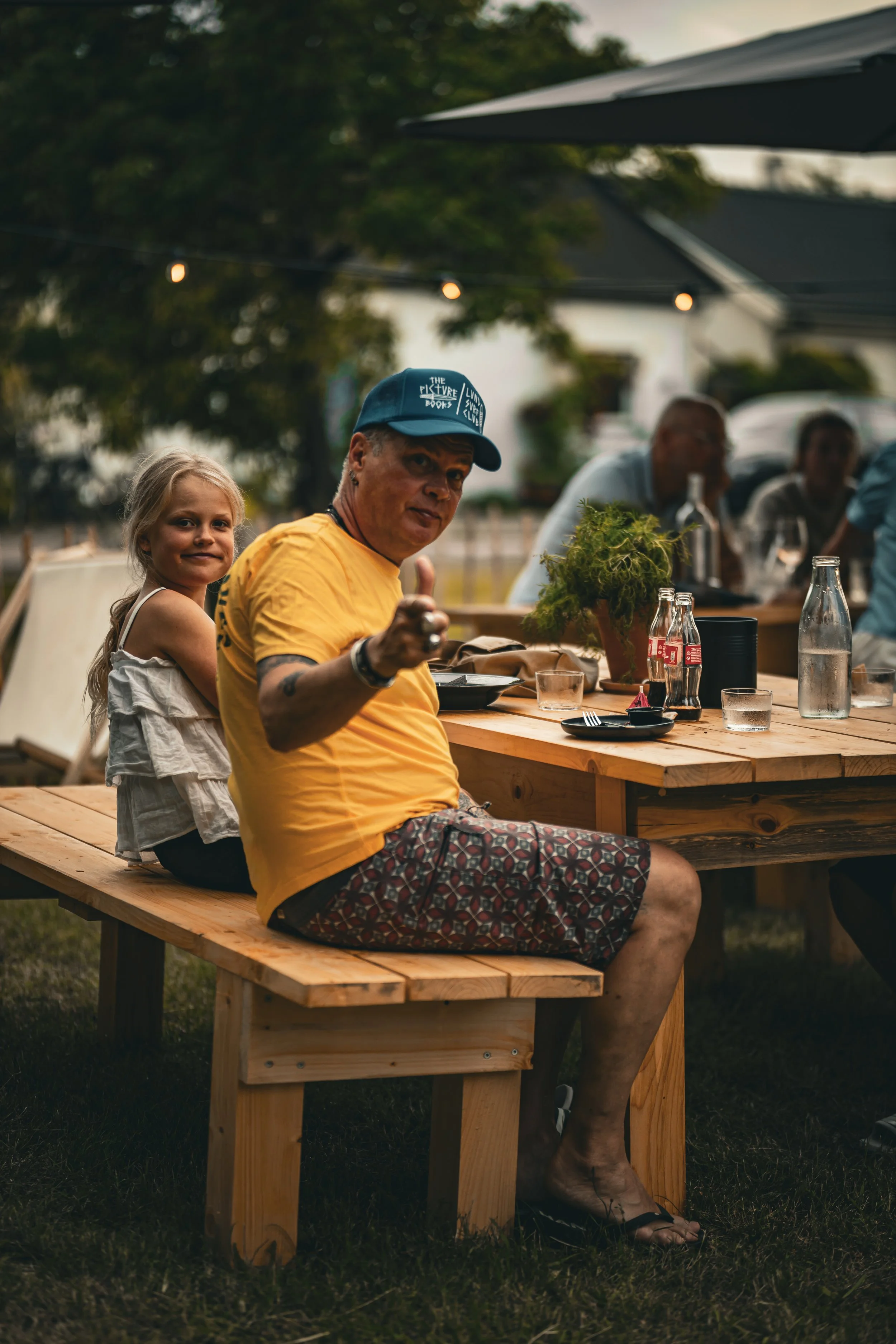 Ung kvinna och man som sitter på en träbänk utomhus vid en fest eller samling. Kvinnan står bakom mannen med armarna runt honom, båda ler och tittar mot kameran. Det finns andra personer i bakgrunden som sitter vid bordet.