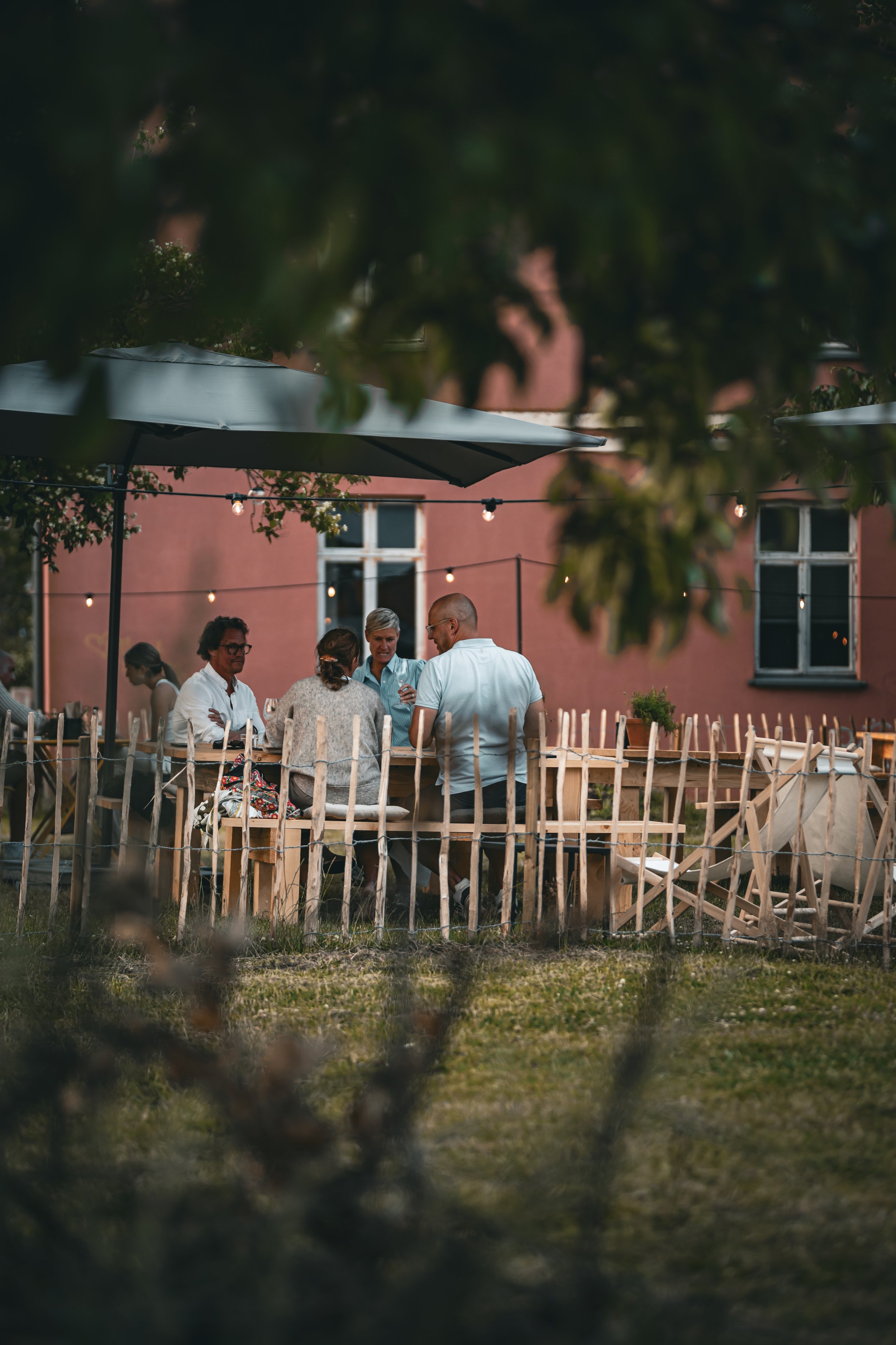 Folk samlade runt ett bord utomhus, omgivna av gröna träd och ett rosa byggnad i bakgrunden.
