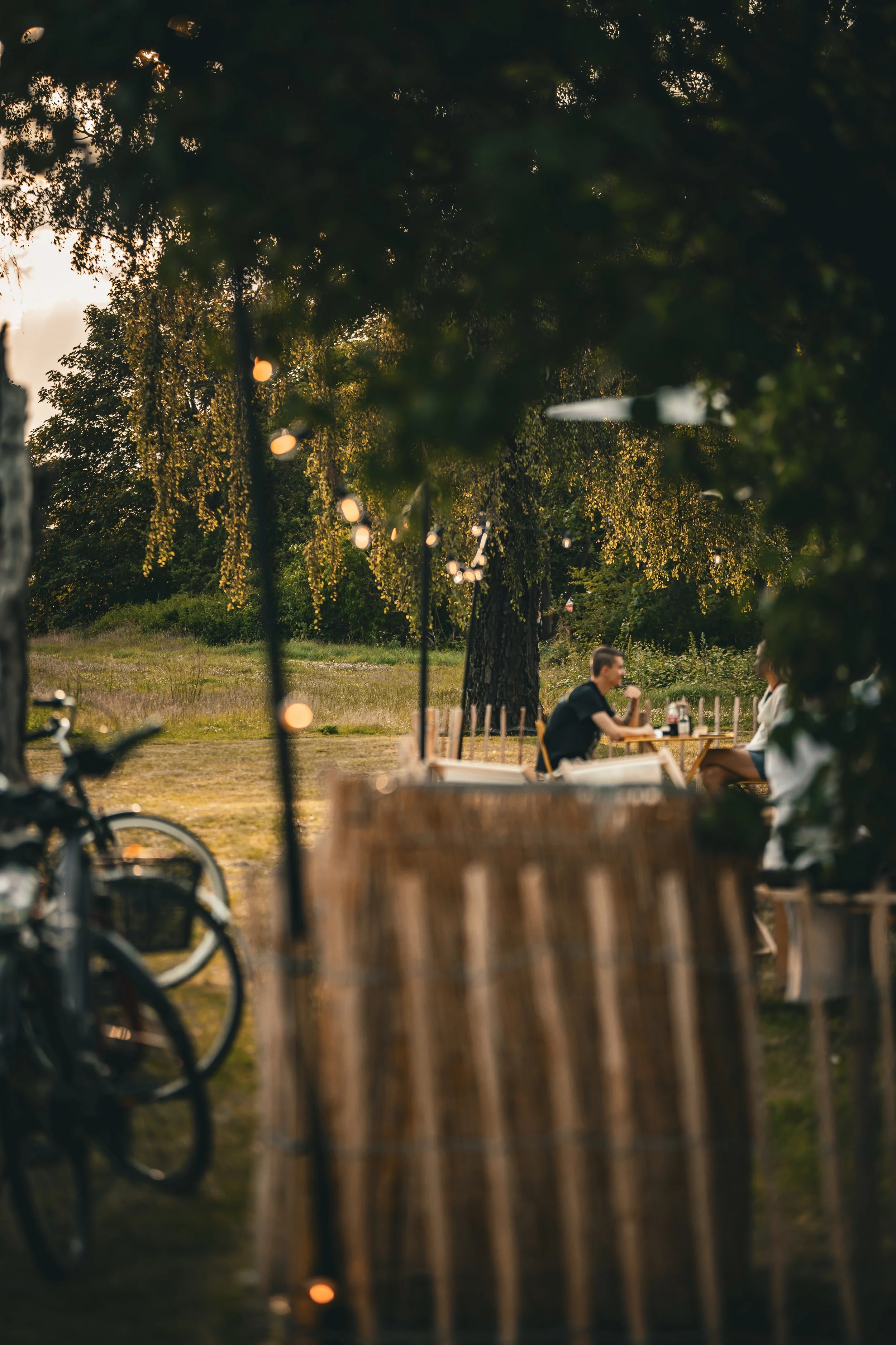 Två personer sitter vid ett dukat utomhusbord, ett träd och lagom kvällsbelysning syns i bakgrunden.