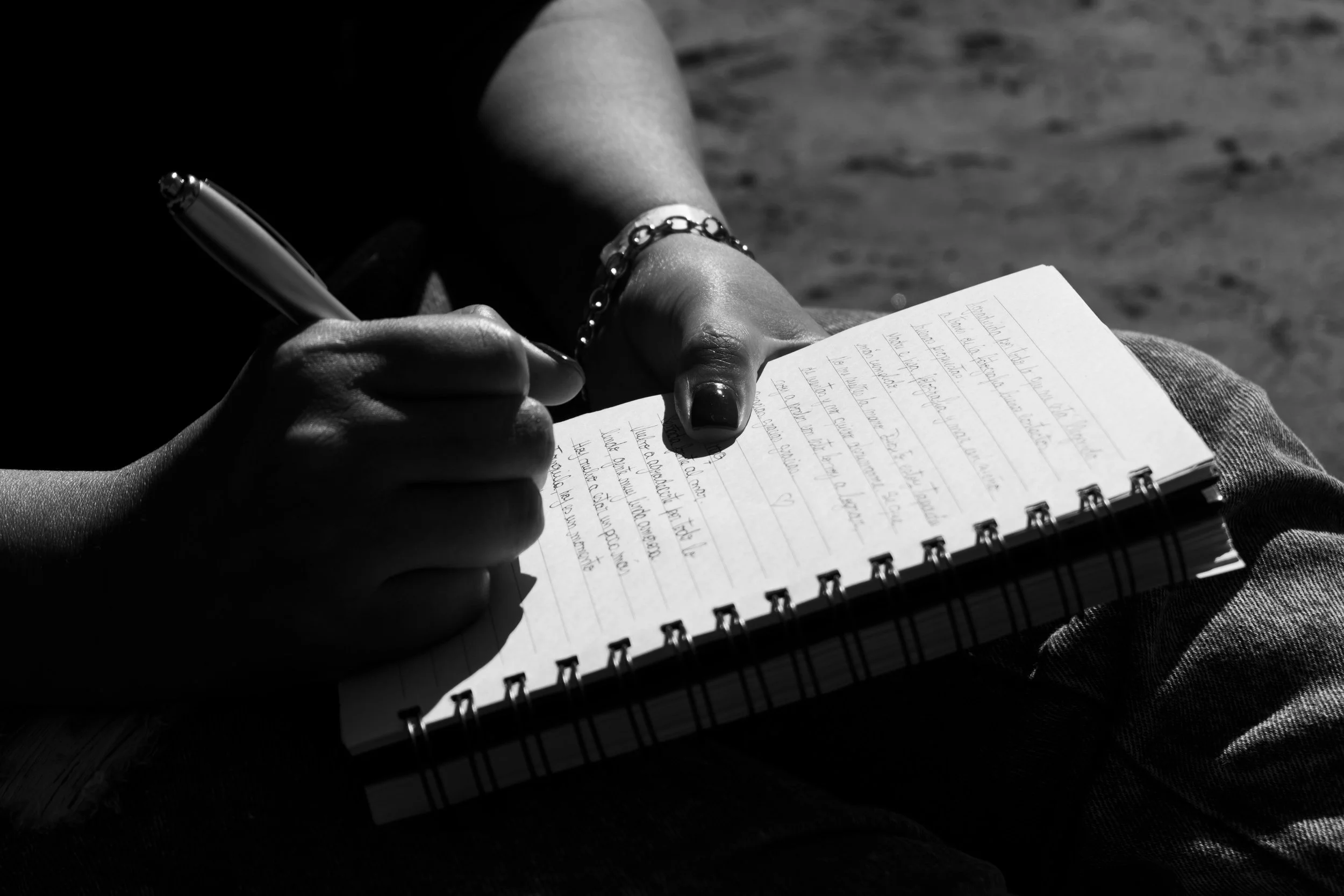 Close-up of a person writing in a spiral notebook with a pen, wearing a bracelet, outdoors on a sunny day.