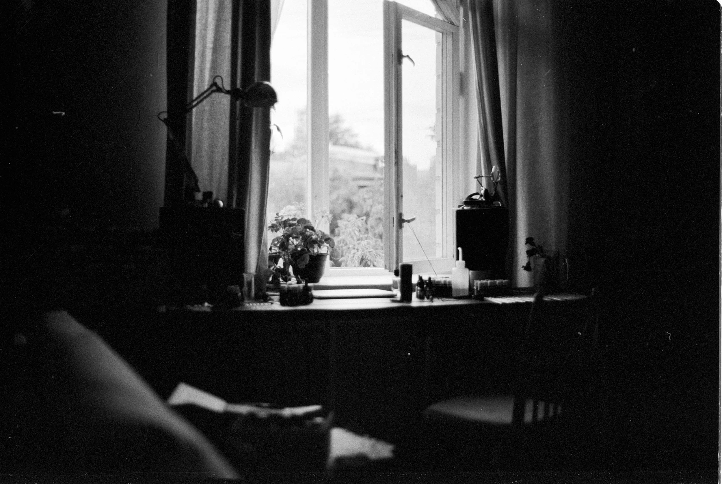 A black-and-white photo of an open window in a room. There are various objects on the windowsill, including potted plants, toiletries, and bottles. Curtains are pulled to the sides, revealing the outside view. The background shows a cloudy sky.