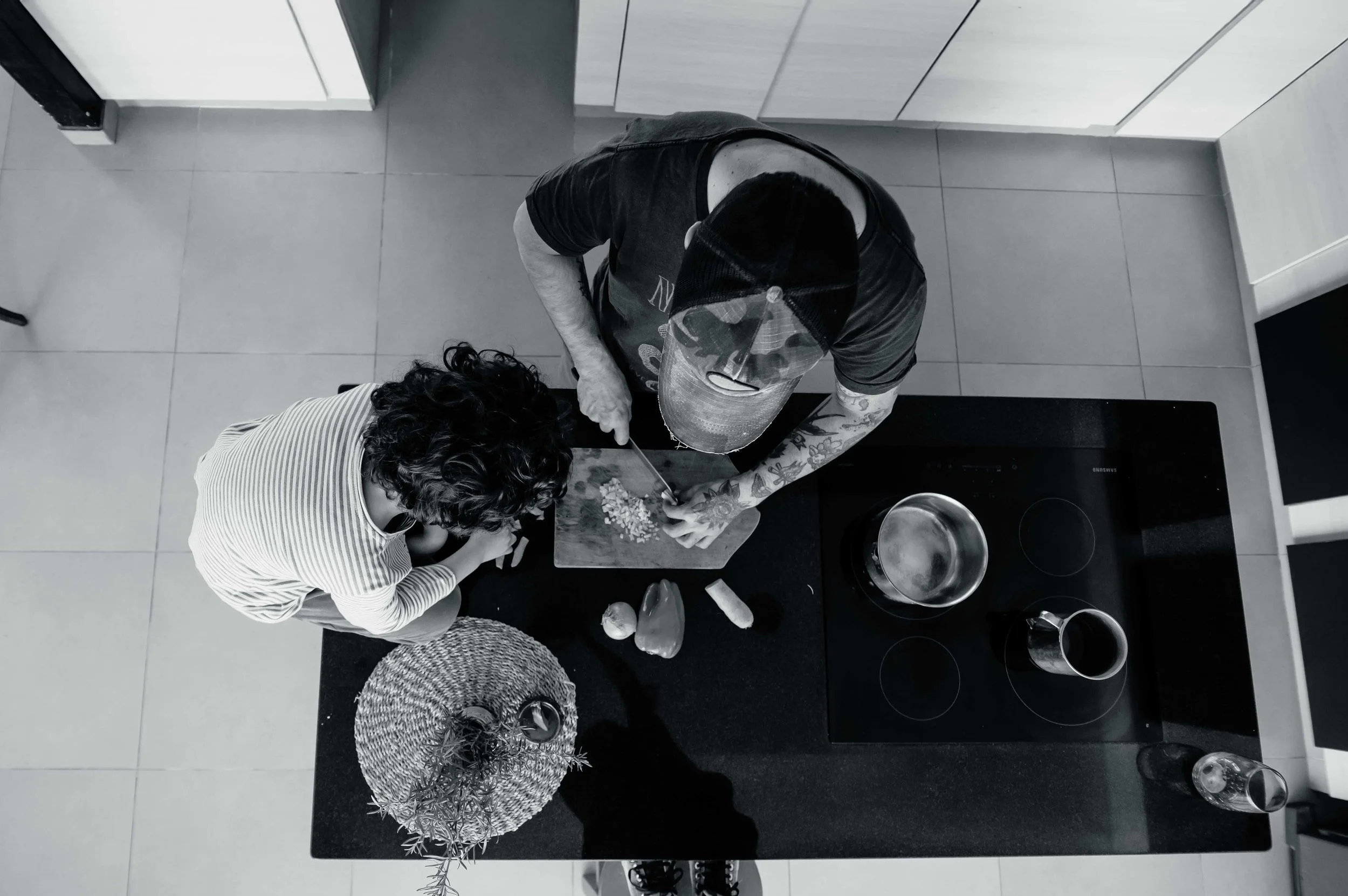 A person with tattoos on their arms and wearing a black cap is chopping vegetables on a cutting board in a kitchen, while another person with curly hair and a striped shirt is watching. There are a pot, a pan, and some other kitchen items on the counter.