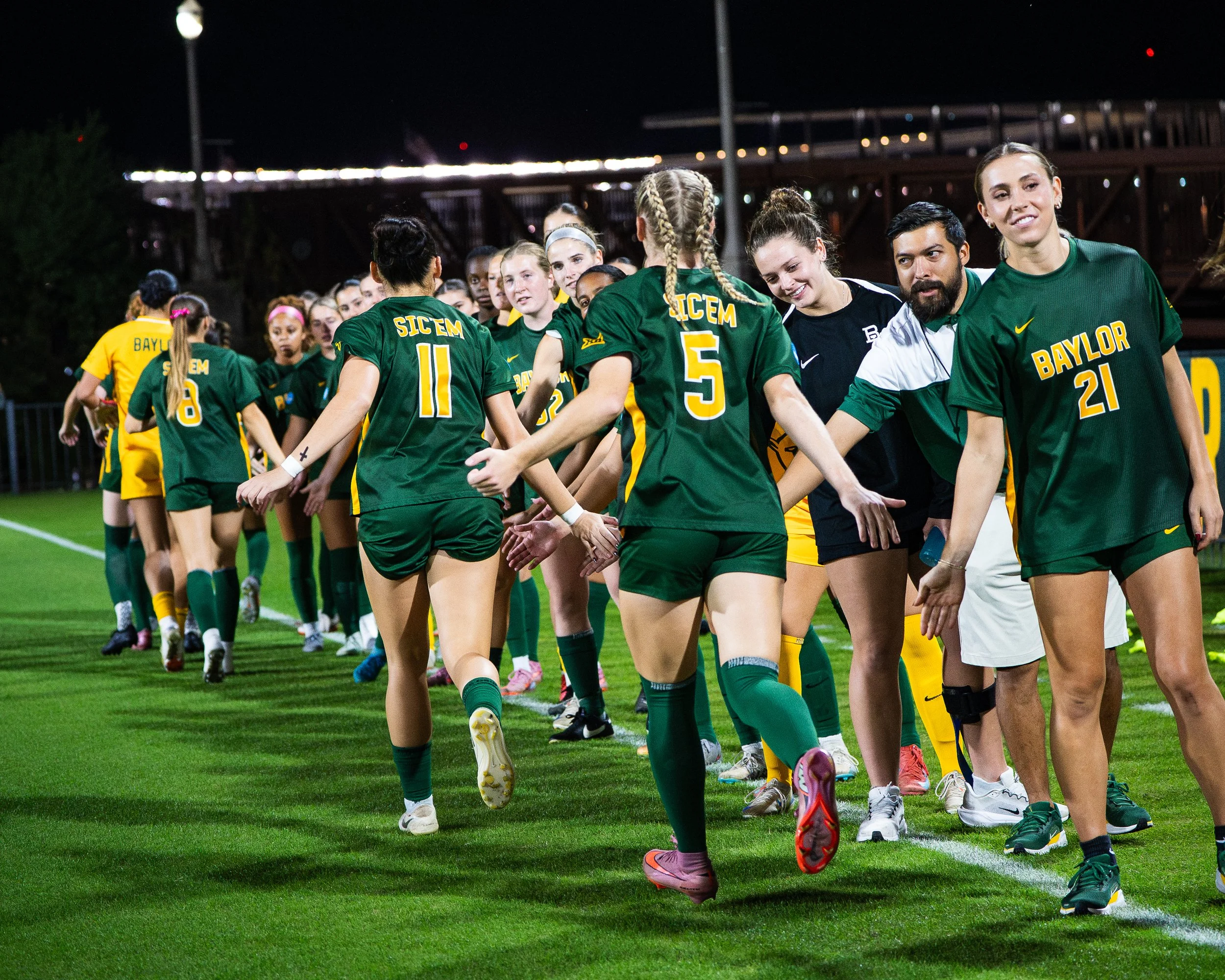 Women’s soccer team wearing green and yellow jerseys shaking hands after a game at night.