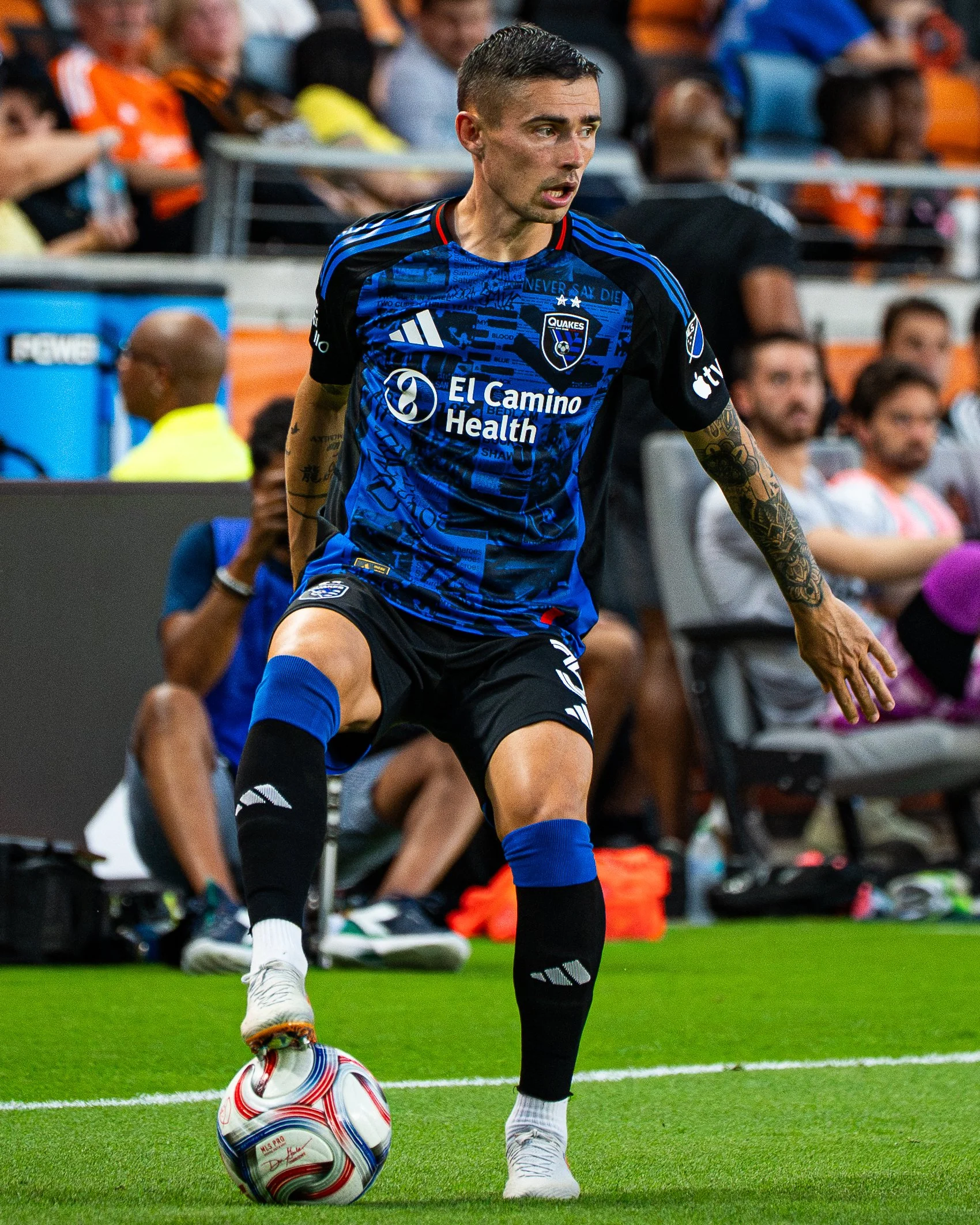 Soccer player in a blue and black uniform about to kick a ball during a match, with spectators in the background.