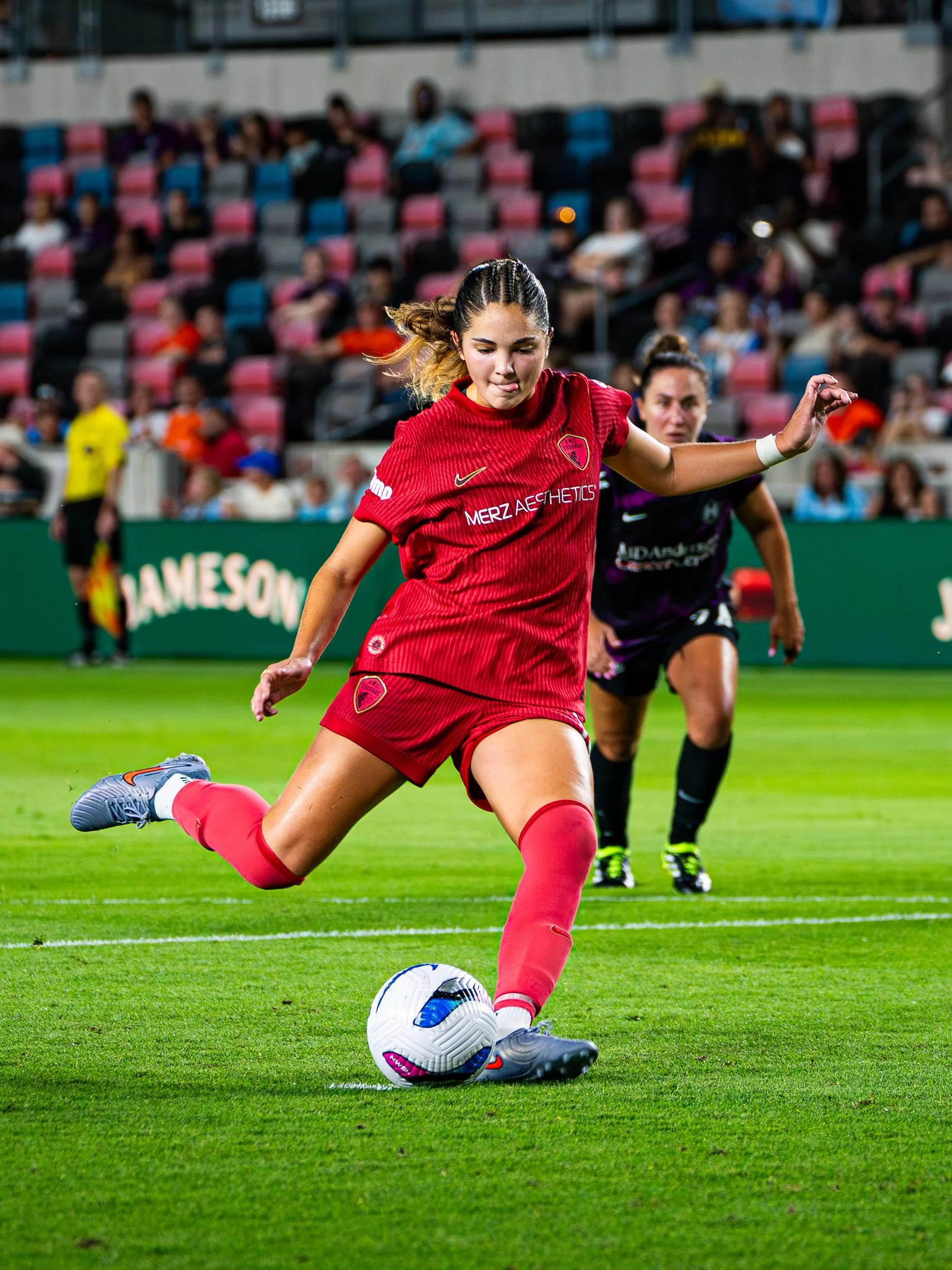 A female soccer player in a red uniform kicking a soccer ball on a green field with spectators in the stands.
