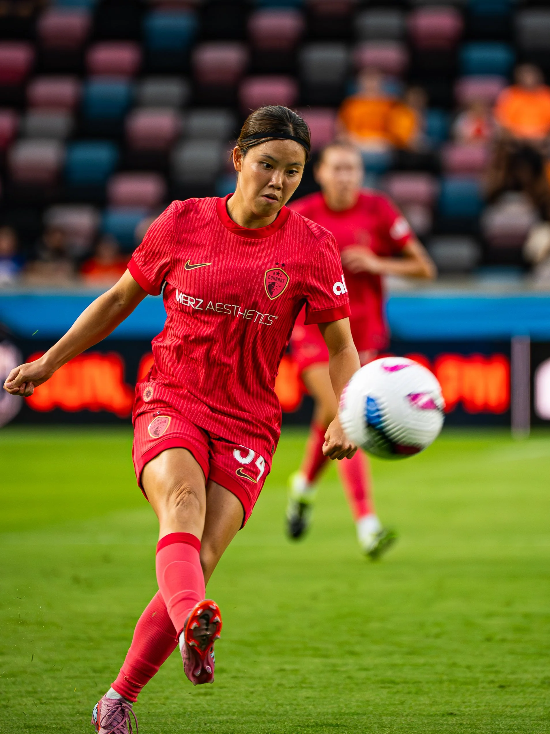 A female soccer player in a red uniform is kicking a soccer ball on a field. Other players and spectators are blurred in the background.