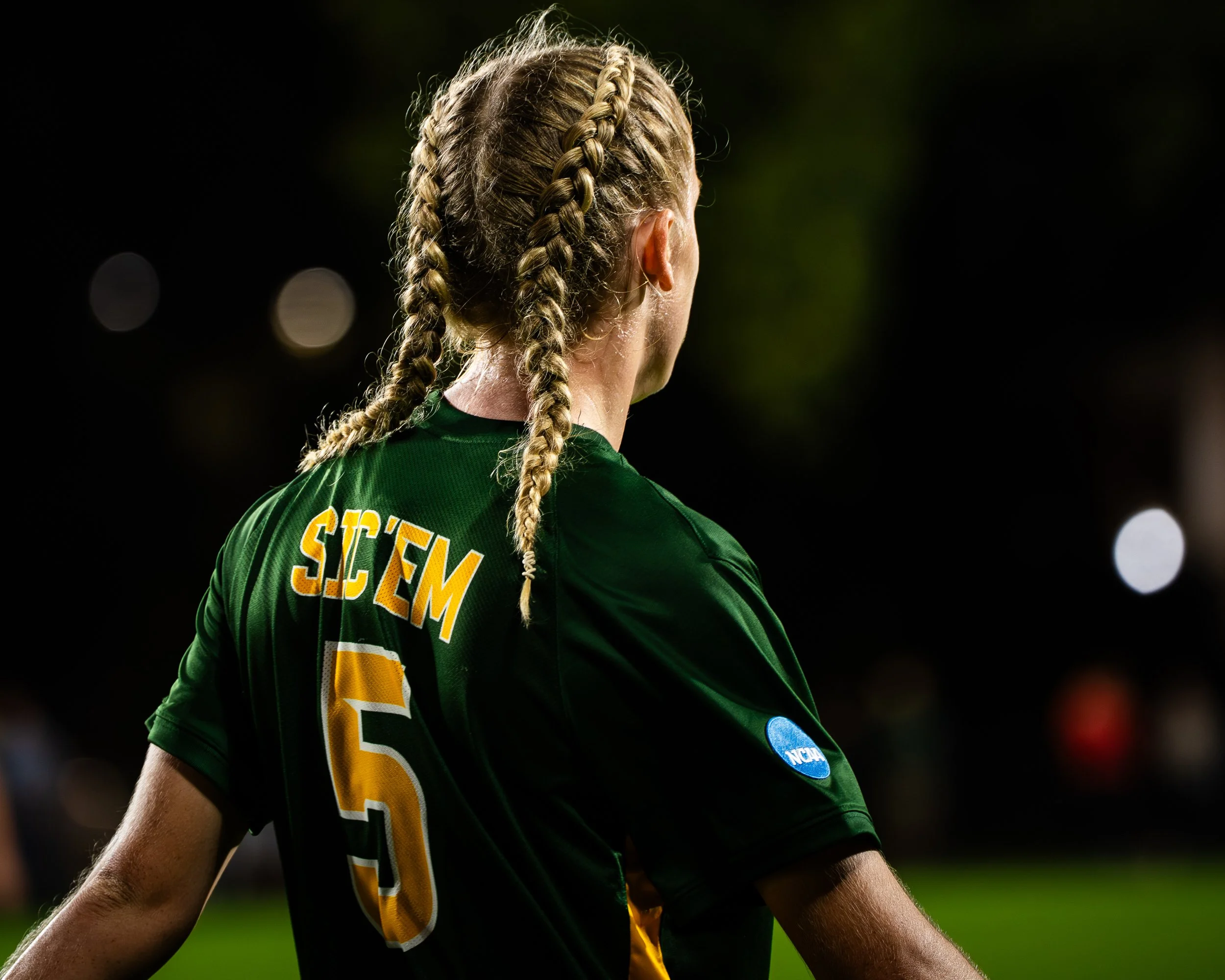 Back view of a female athlete with blonde braids wearing a green sports jersey with yellow and white lettering, and a blue NCAA sticker.