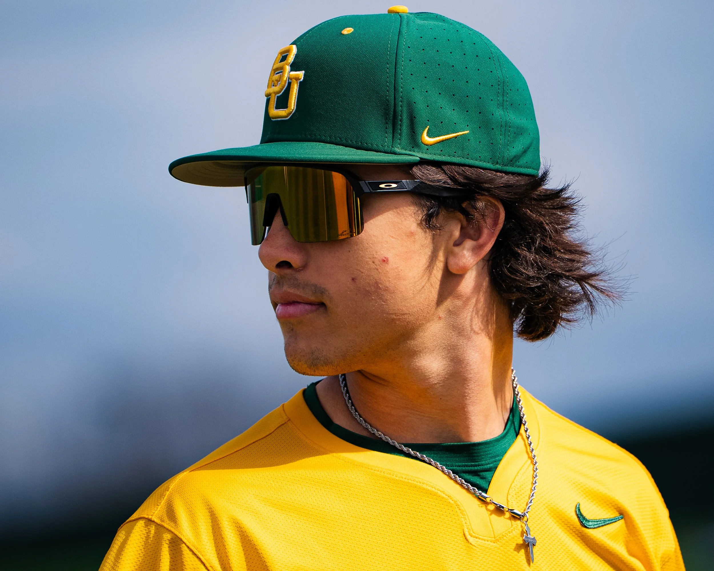 Close-up of a young man wearing a green baseball cap with a yellow logo, reflective sports sunglasses, and a yellow sports jersey with green details, against a blue sky background.