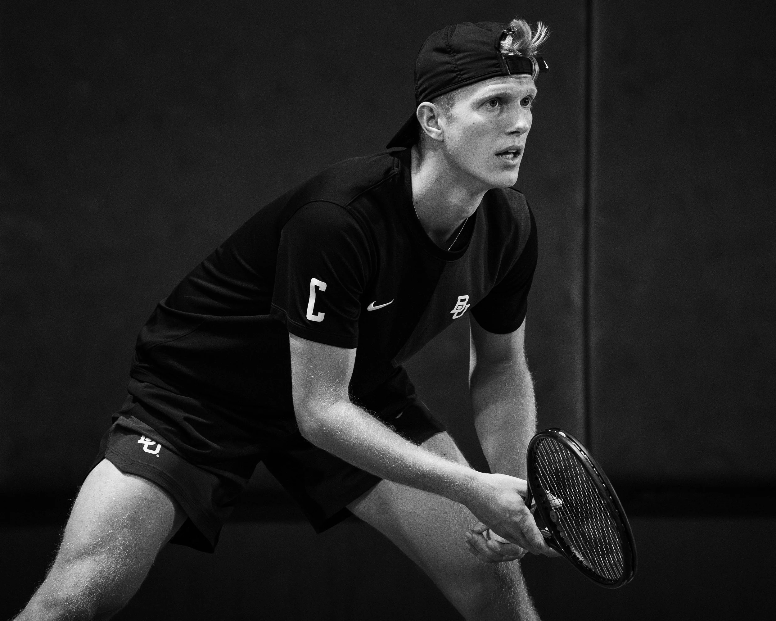 A young man playing tennis indoors, wearing a black cap backward, a black sports shirt, and holding a tennis racket, with a focused expression.