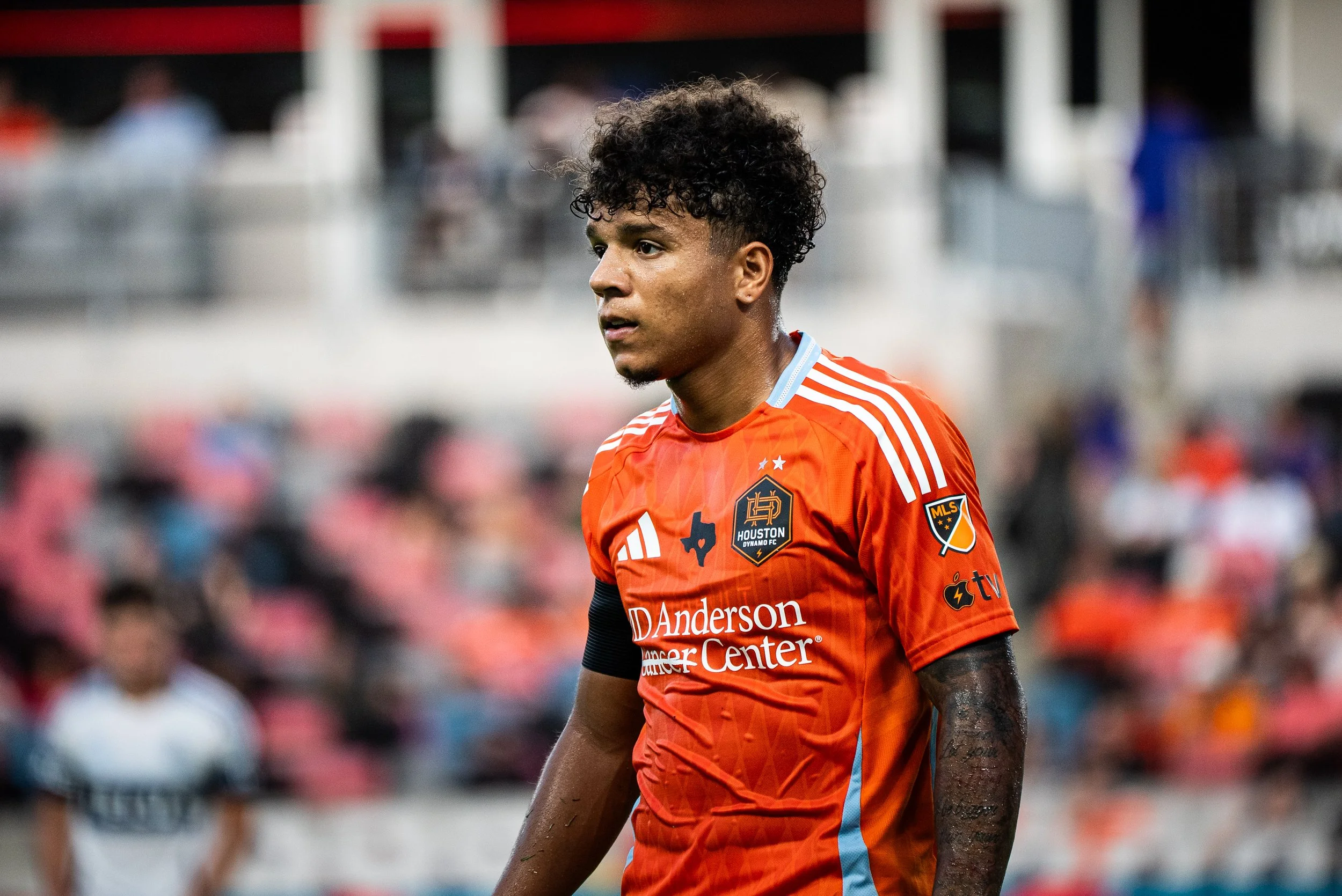 A soccer player wearing an orange Houston Dynamo FC jersey with sponsorship logos stands on the field during a match, with blurred spectators in the background.
