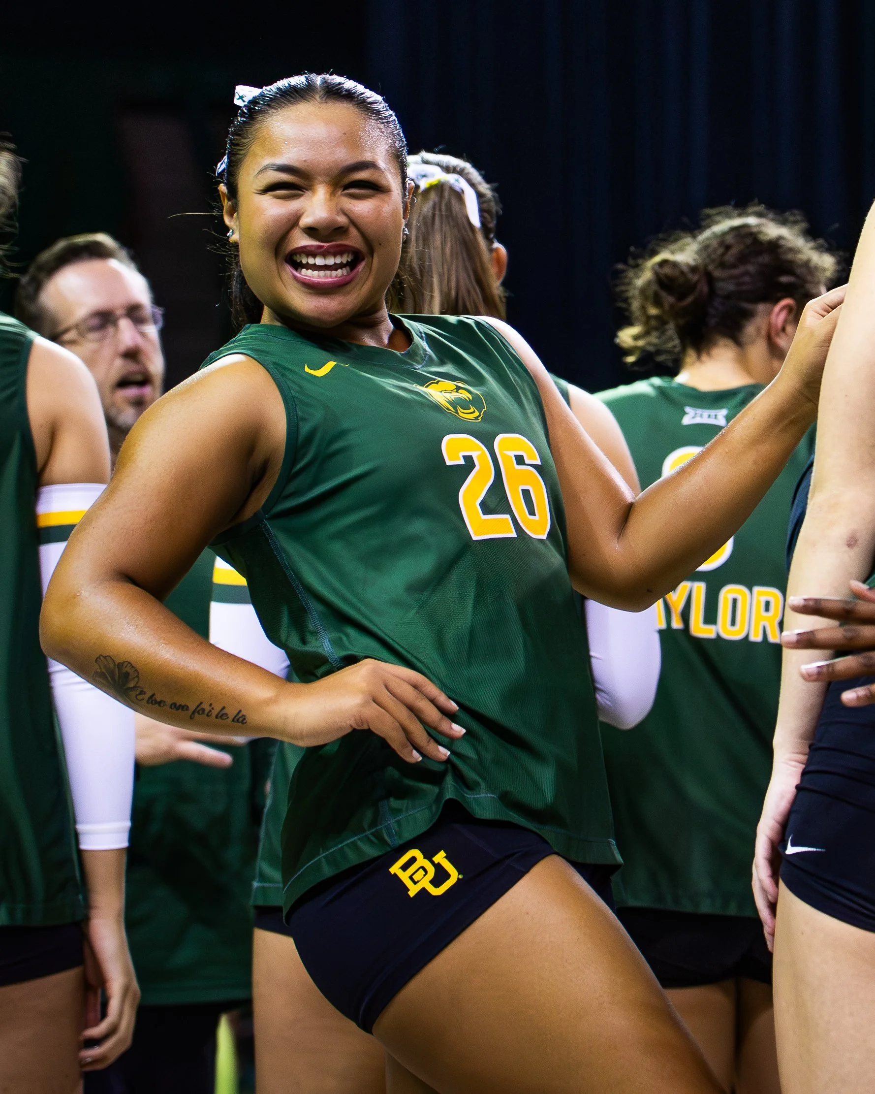 Close-up of an Asian female volleyball player wearing a green jersey with the number 26, smiling, with other players around her. The background is dark, and she is standing with her hand on her hip.