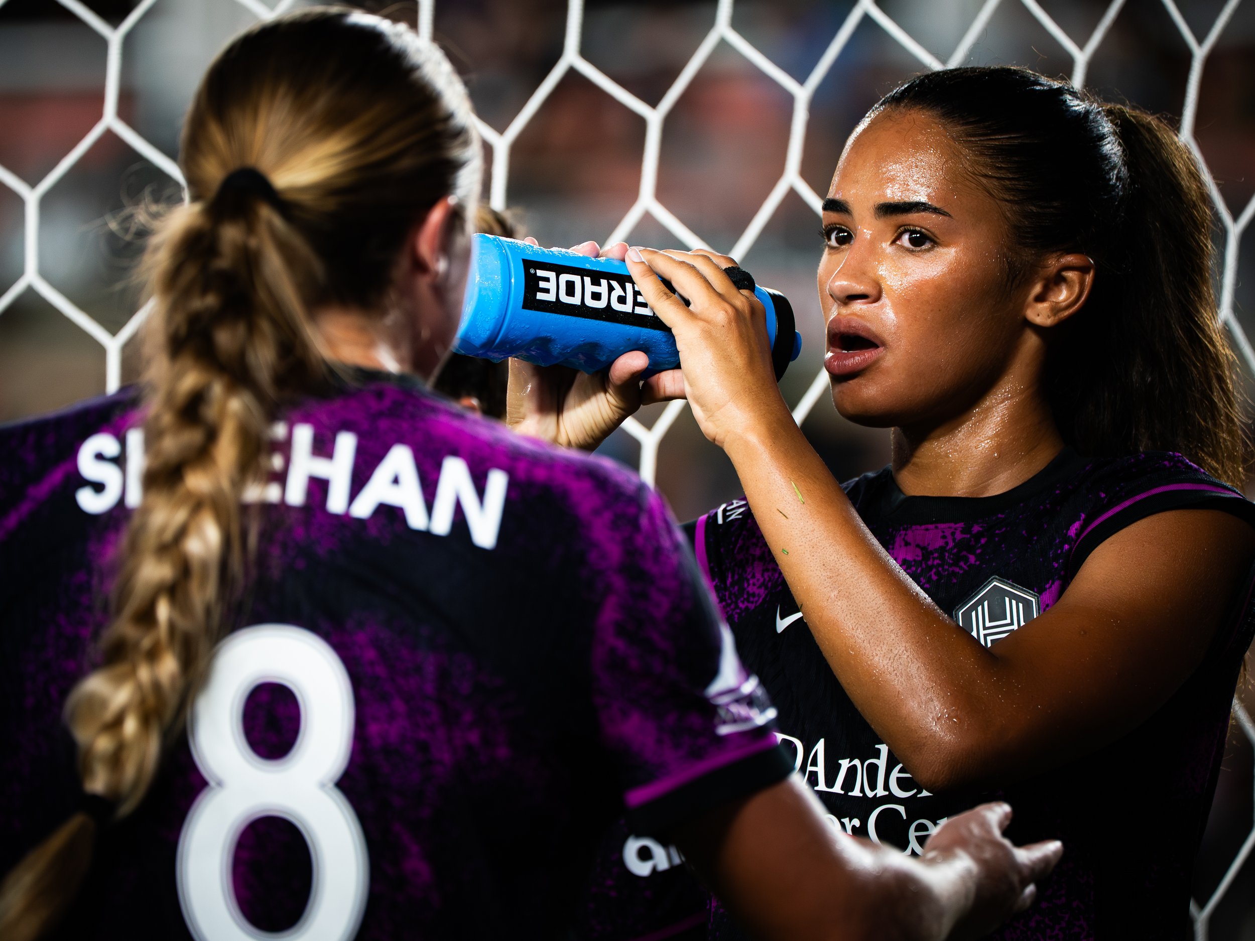 Two female soccer players, one with long braided hair and a jersey that reads 'SHEHAN' and number 8, and the other with wet hair, are close to each other by the goal net. The player with wet hair is holding a blue water bottle near her mouth.