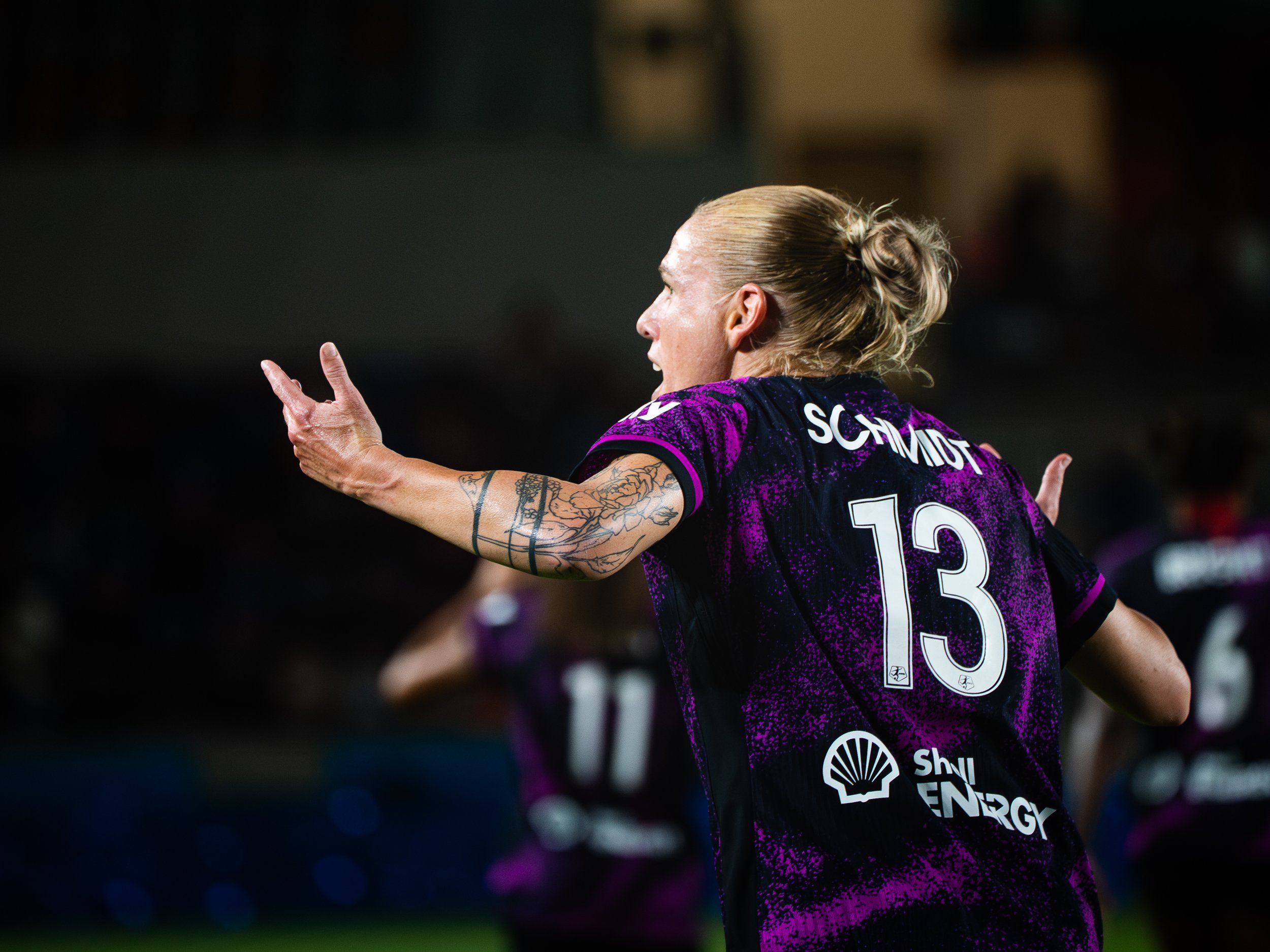A female soccer player with blonde hair in a bun, tattoos on her arm, wearing a purple and black jersey with the number 13, celebrating on the field with her arms outstretched.
