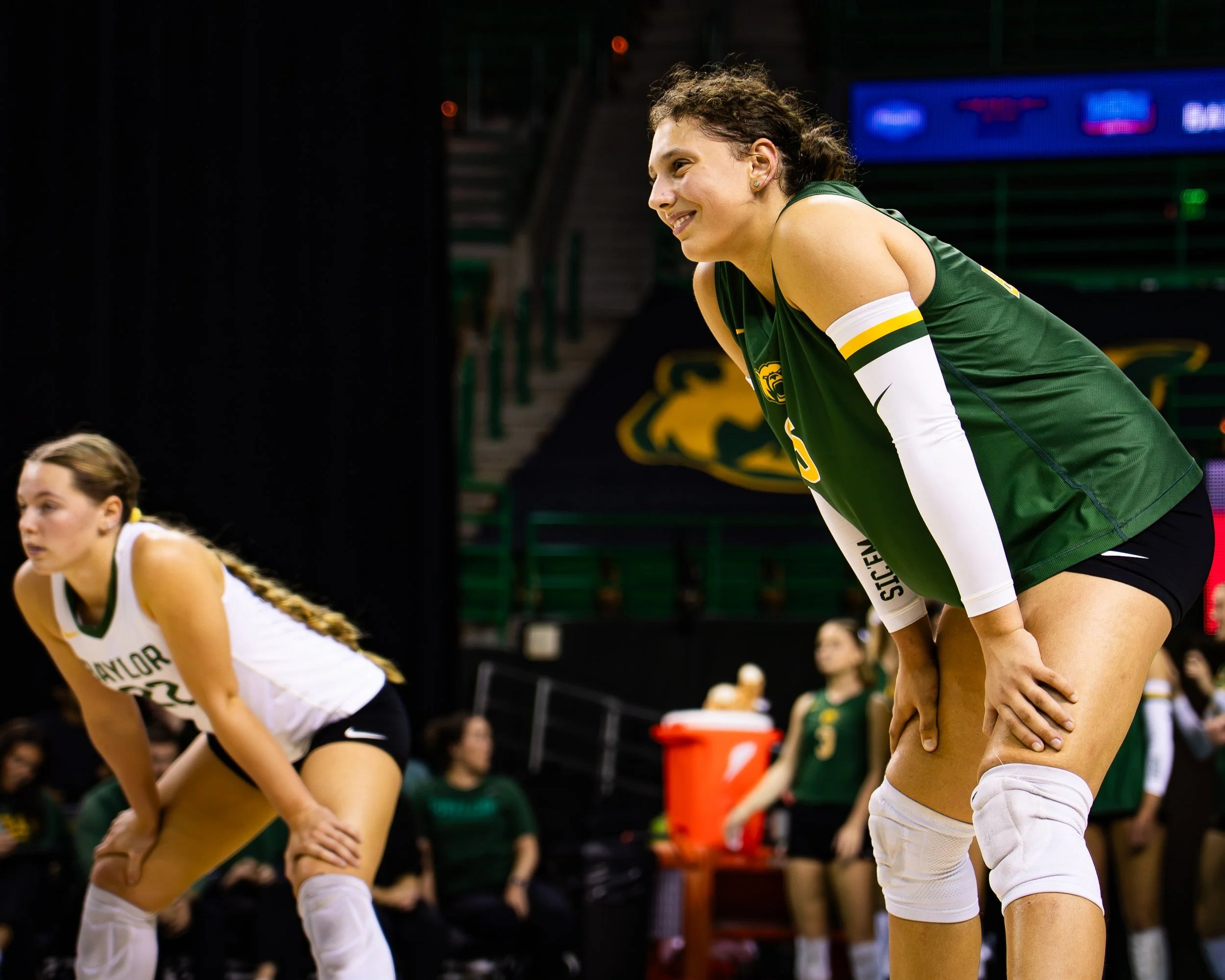 Two female volleyball players wearing green and white uniforms are bending over with hands on their knees during a game, with a gymnasium in the background.
