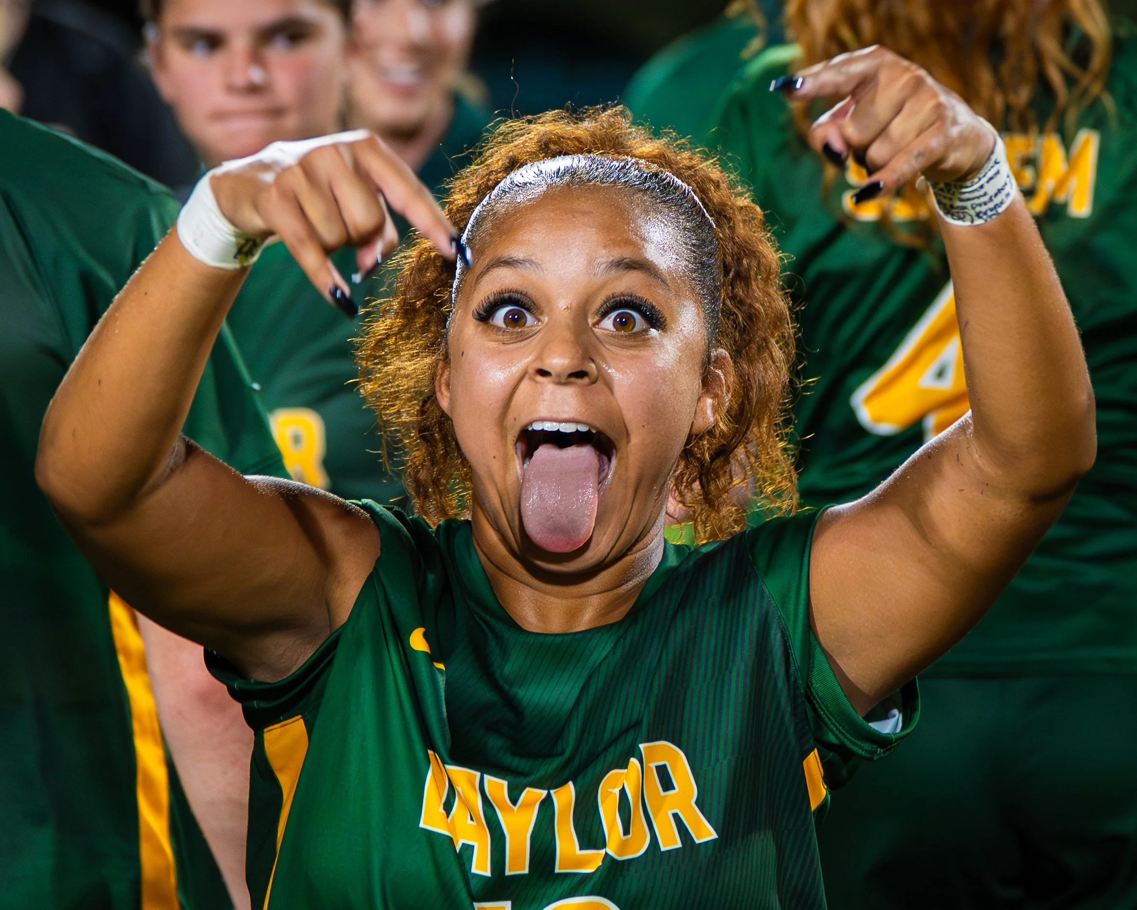 A woman with curly red hair sticking her tongue out and making a rock sign with her hand, surrounded by other women in green sports uniforms in a sporting event.