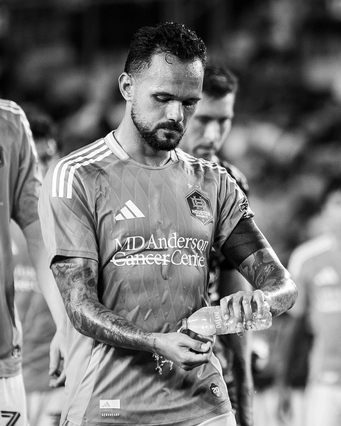 A soccer player with tattoos on his arms, wearing a Houston Dynamo jersey, holding a water bottle, and looking down.