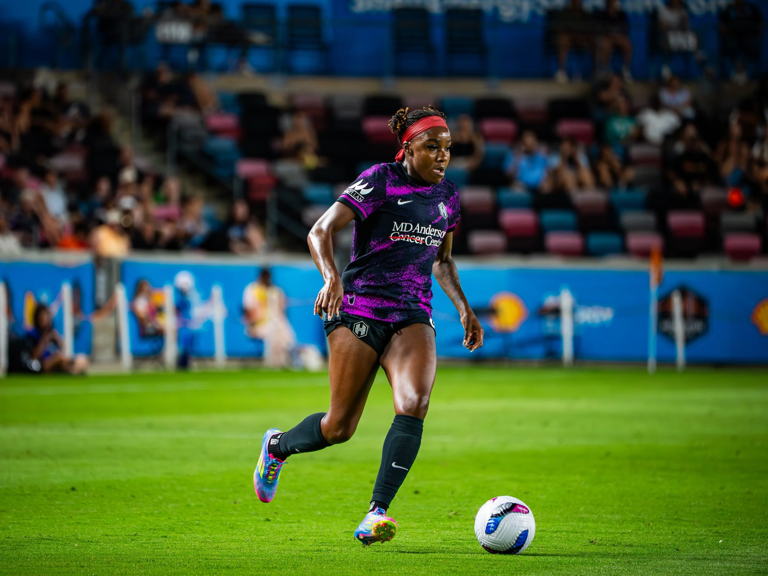 A female soccer player in a purple and black uniform with a red headband is kicking a soccer ball on a field during a game, with spectators in the background.