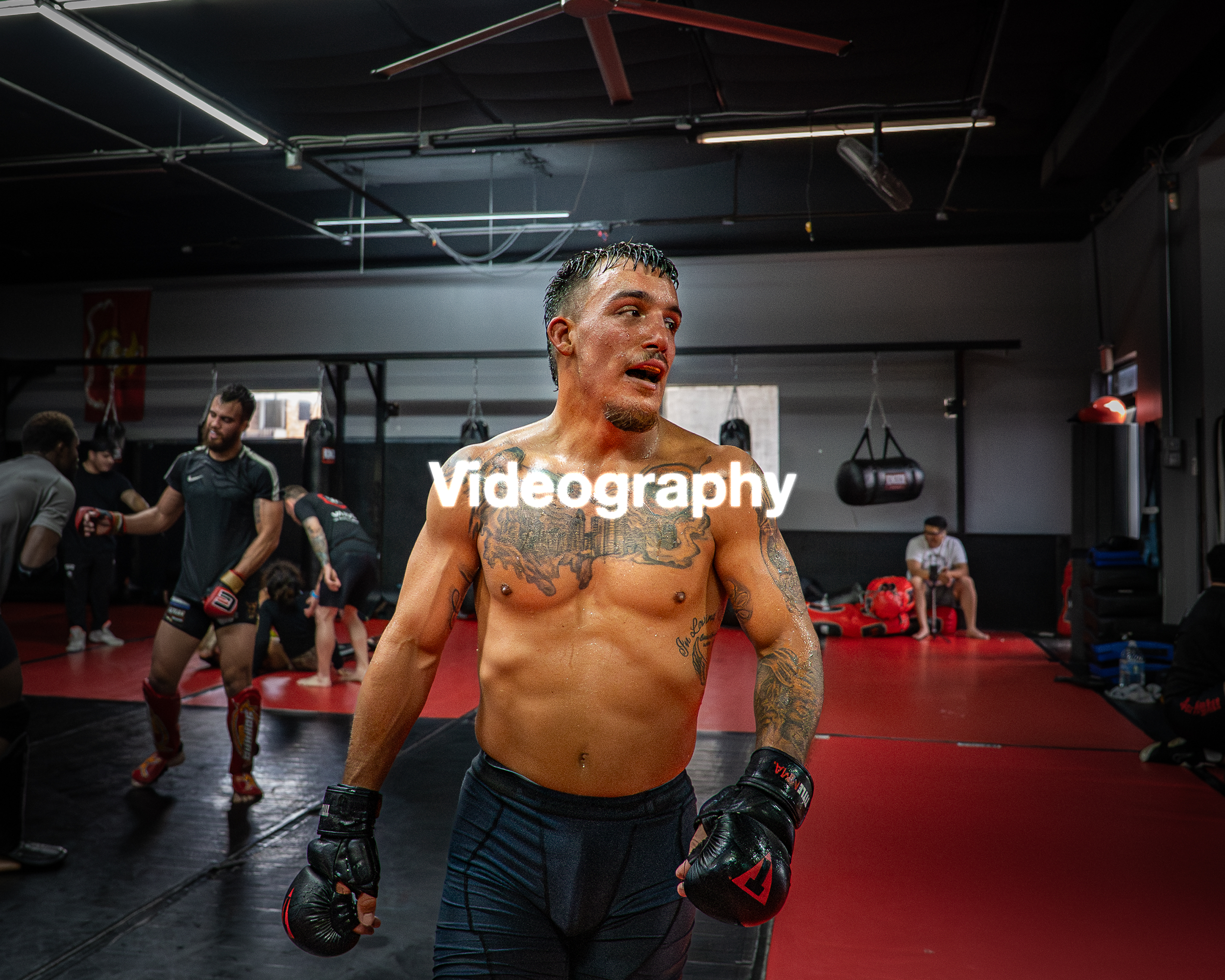 A shirtless male boxer with tattoos is standing in a boxing gym, holding boxing gloves and a mouthguard, with other fighters and trainers in the background preparing for training or sparring.
