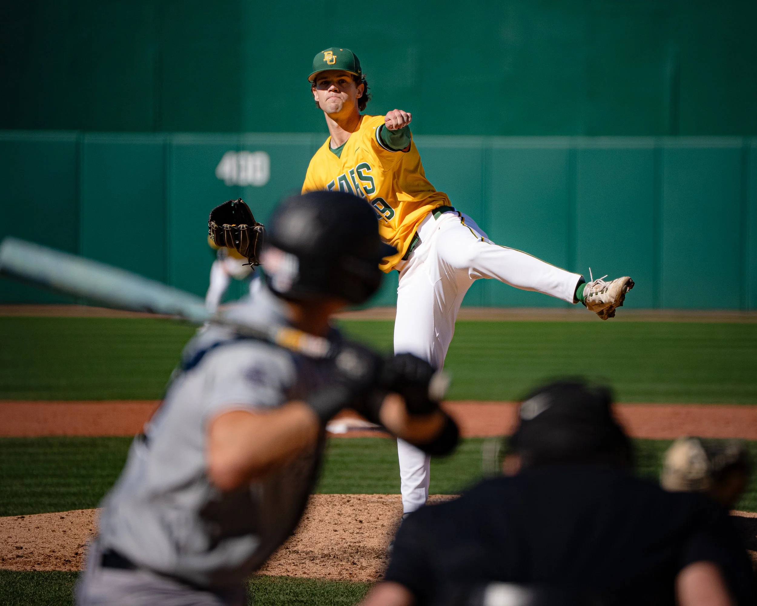 A baseball pitcher in a yellow jersey and green cap throwing a pitch, with players in white uniforms on the field.