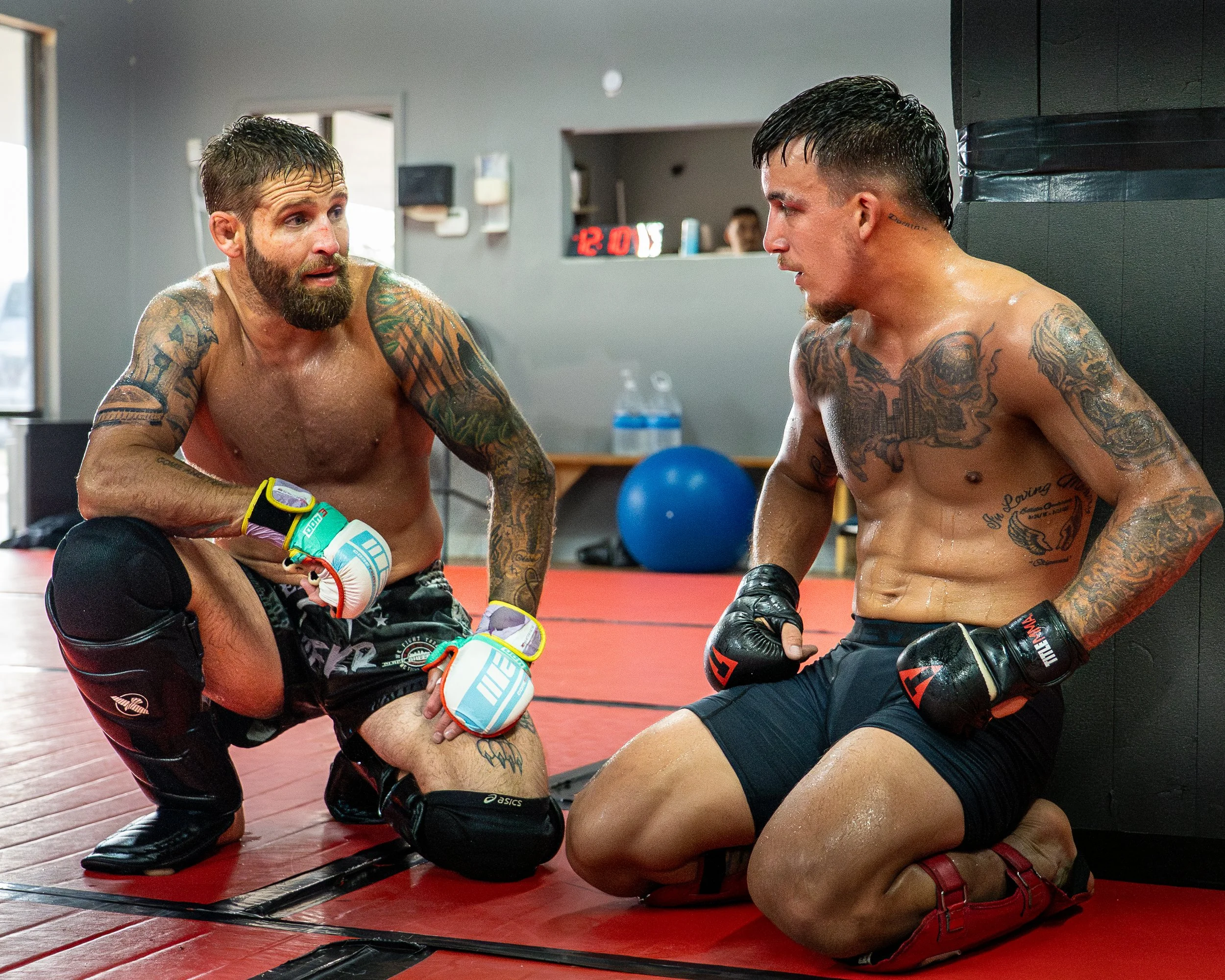 Two shirtless male mixed martial artists with tattoos, wearing boxing gloves and shorts, are engaged in a fight inside a gym. One is kneeling while the other is leaning against the wall. The scene appears intense with sweat visible on their bodies.