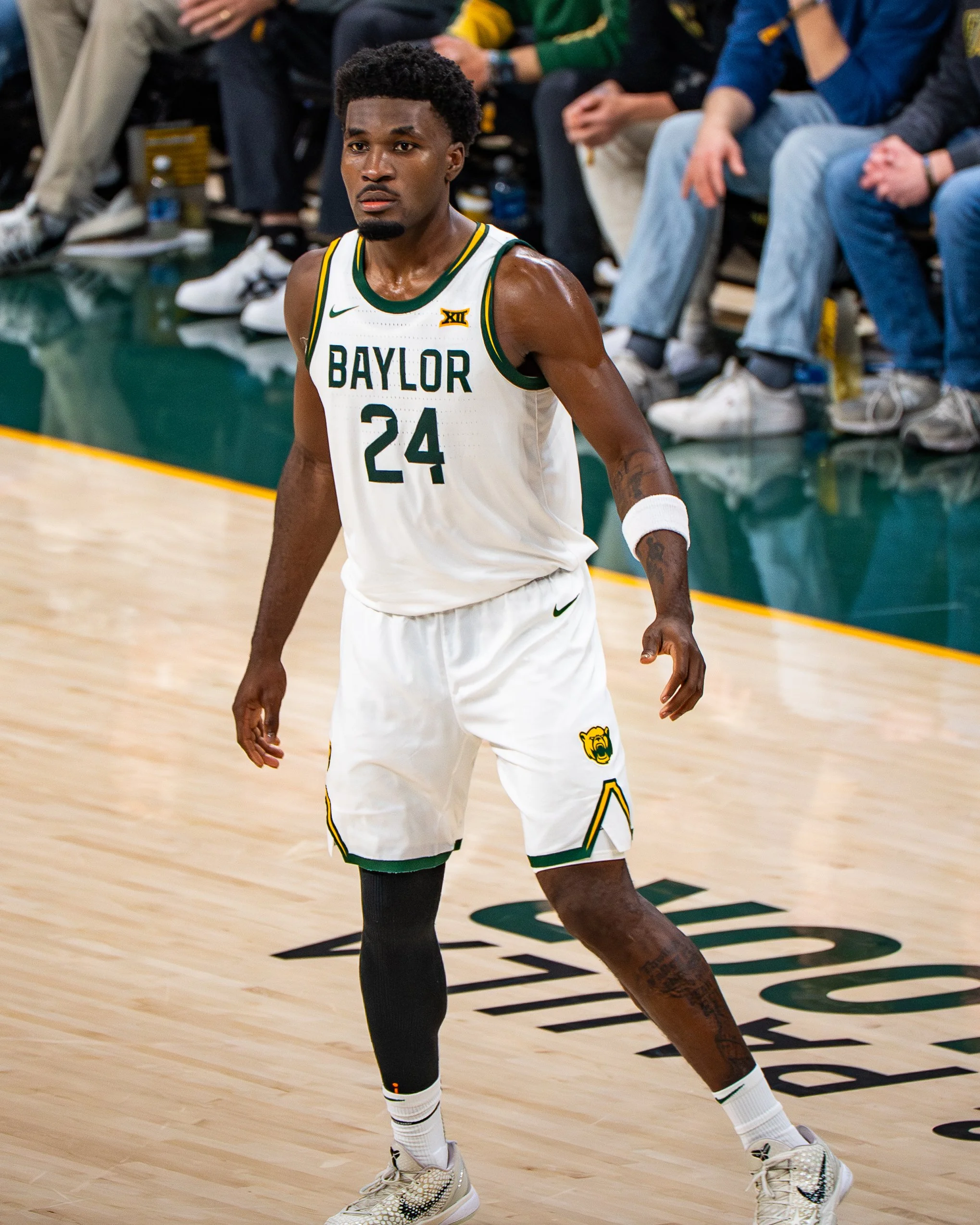 A male basketball player in a Baylor basketball uniform with the number 24, standing on the court during a game, with spectators seated in the background.